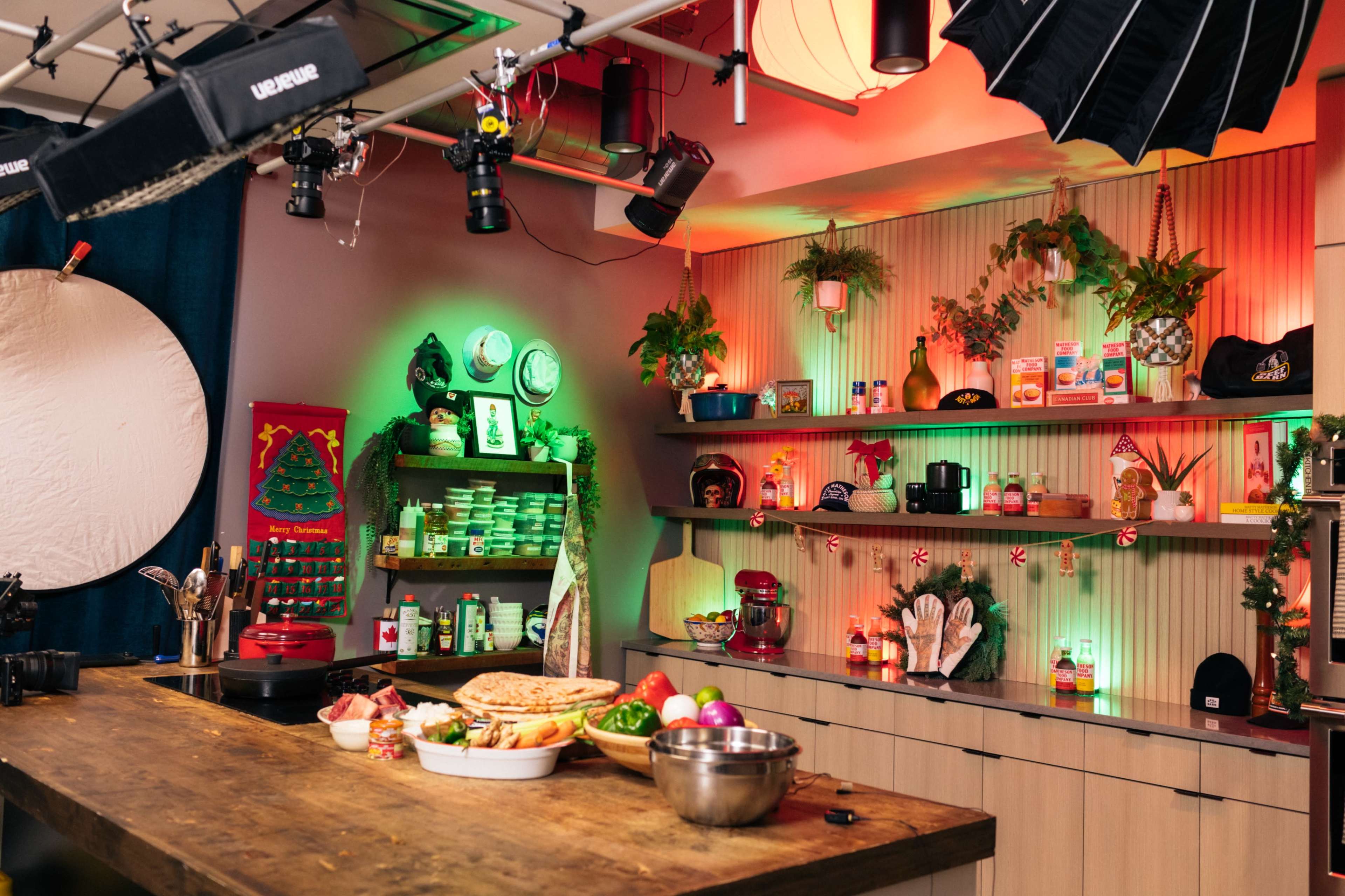 A brightly lit kitchen set features a wooden countertop with various ingredients, pots, and a mixer, surrounded by shelves decorated with colorful jars and plants.
