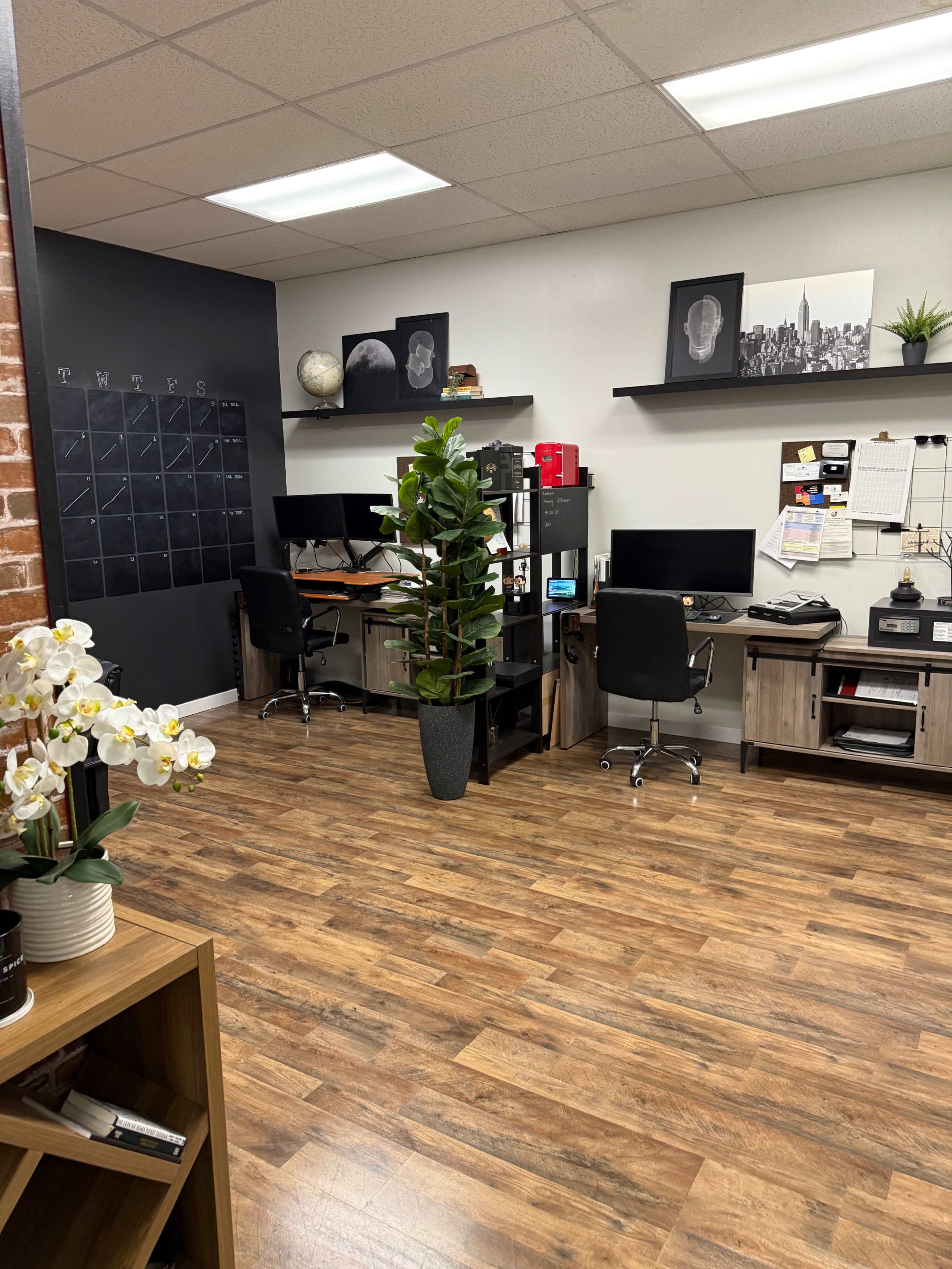 The image shows a modern office space with two desks, a decorative plant, and a calendar on the wall.