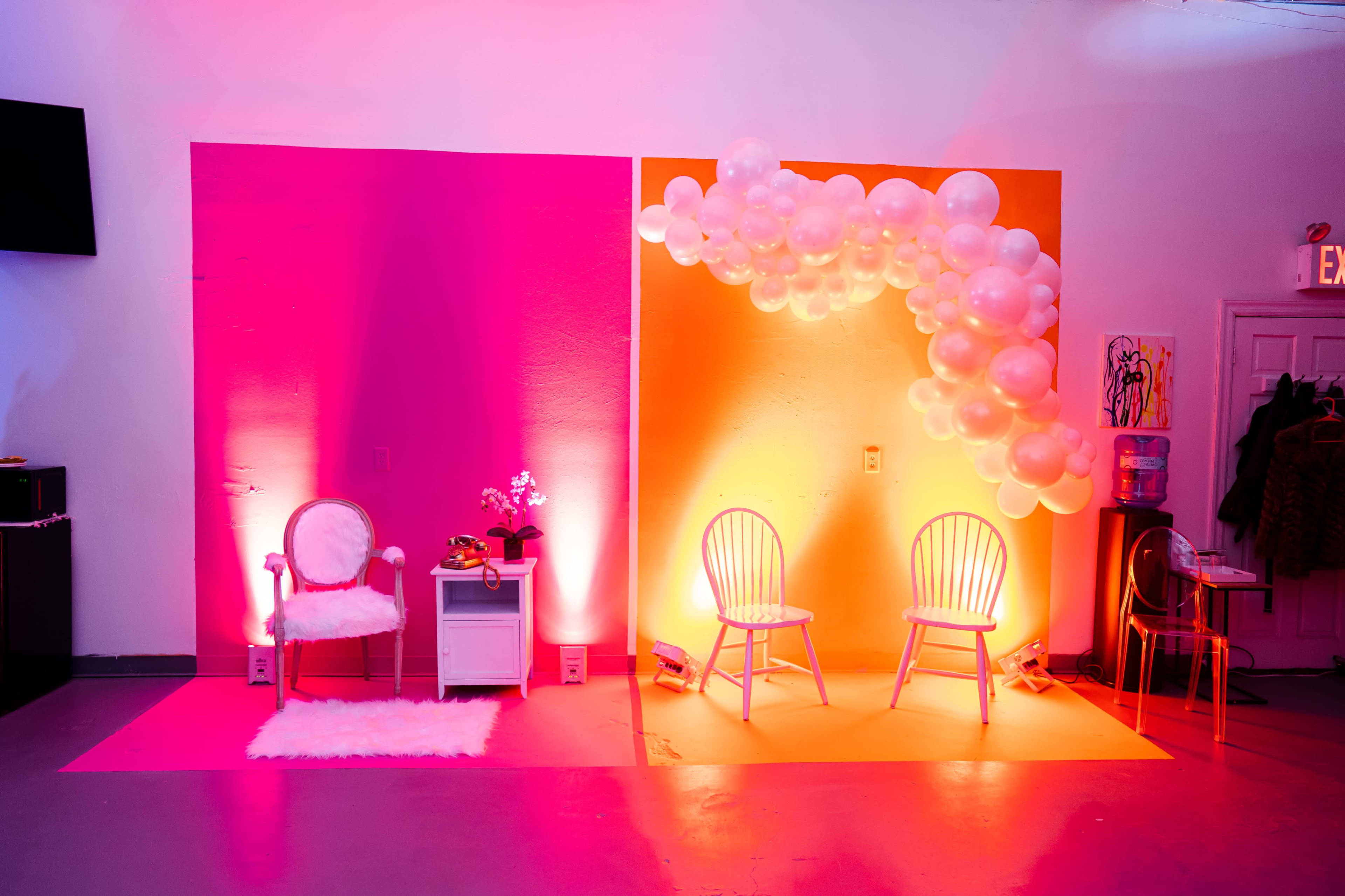 The scene features a brightly lit event space divided by two colored backdrops, one pink and one orange, with decorative balloons and two chairs set up on each side.