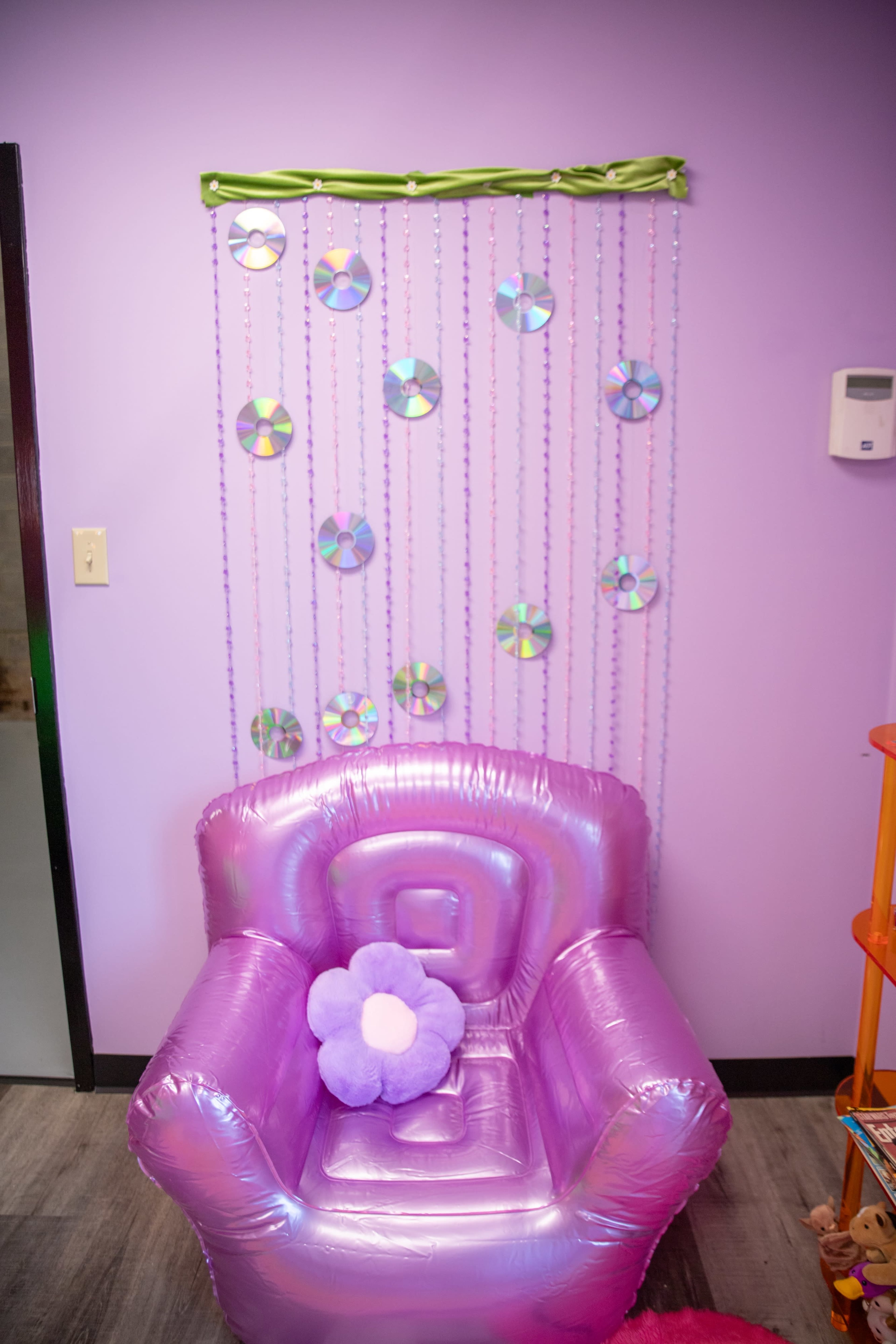 The image shows a purple children's room featuring a shiny inflatable chair and a wall adorned with hanging decorations made of CDs and beads.