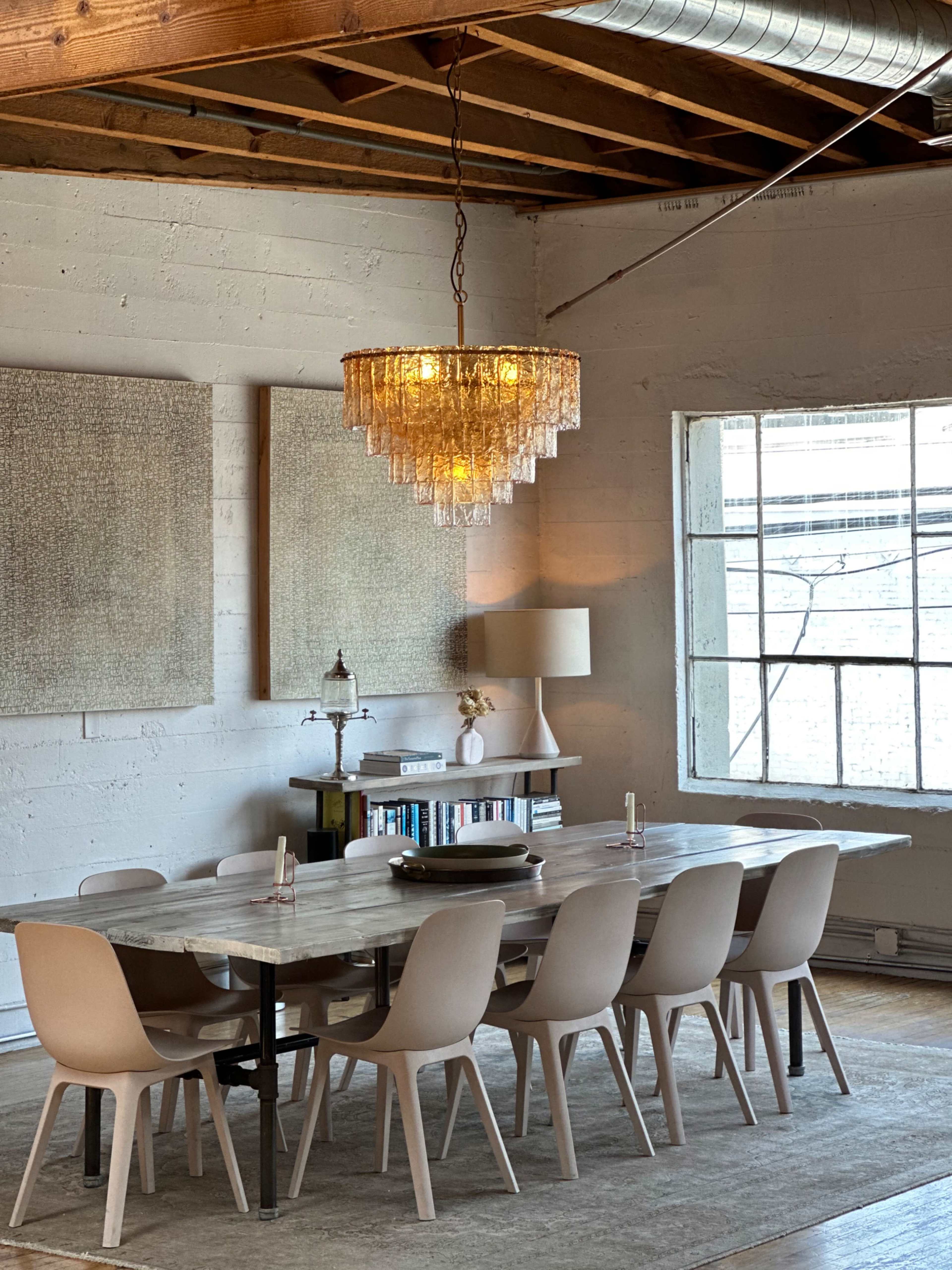 A spacious dining room features a long wooden table surrounded by ten minimalist chairs, a crystal chandelier hangs above, and large windows allow natural light to fill the space.