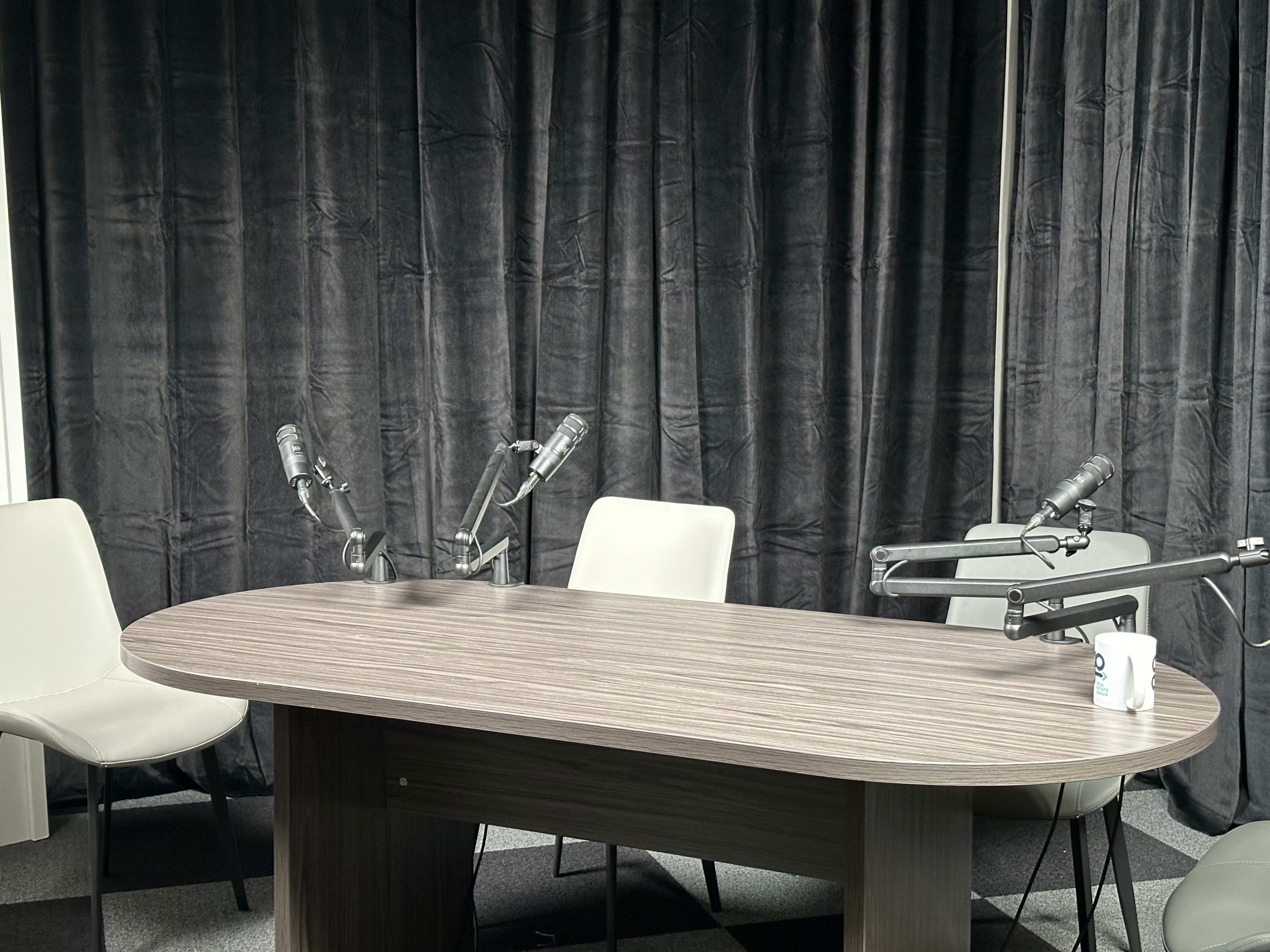 A round wooden table is set in a recording studio with three chairs and microphones positioned around it, against a backdrop of black curtains.