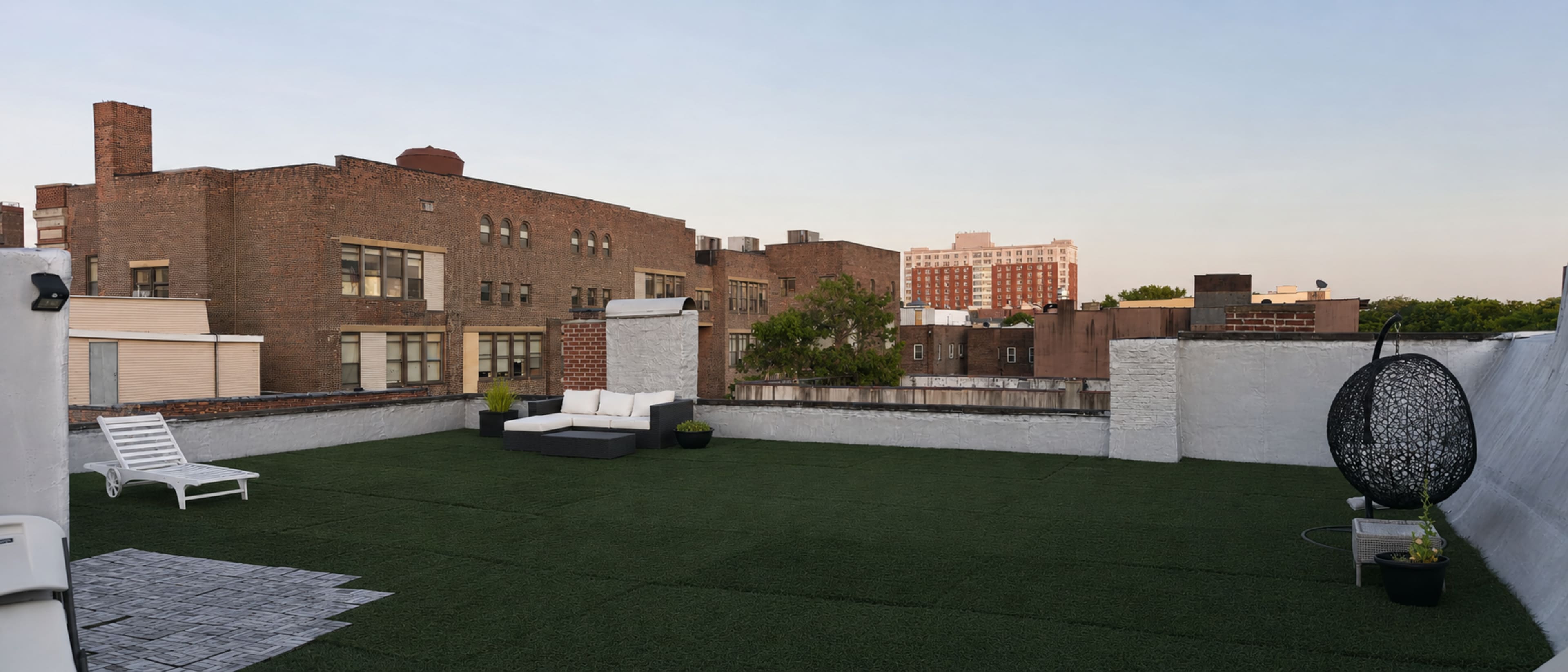The image shows a rooftop terrace featuring artificial grass, modern seating, and views of surrounding buildings.
