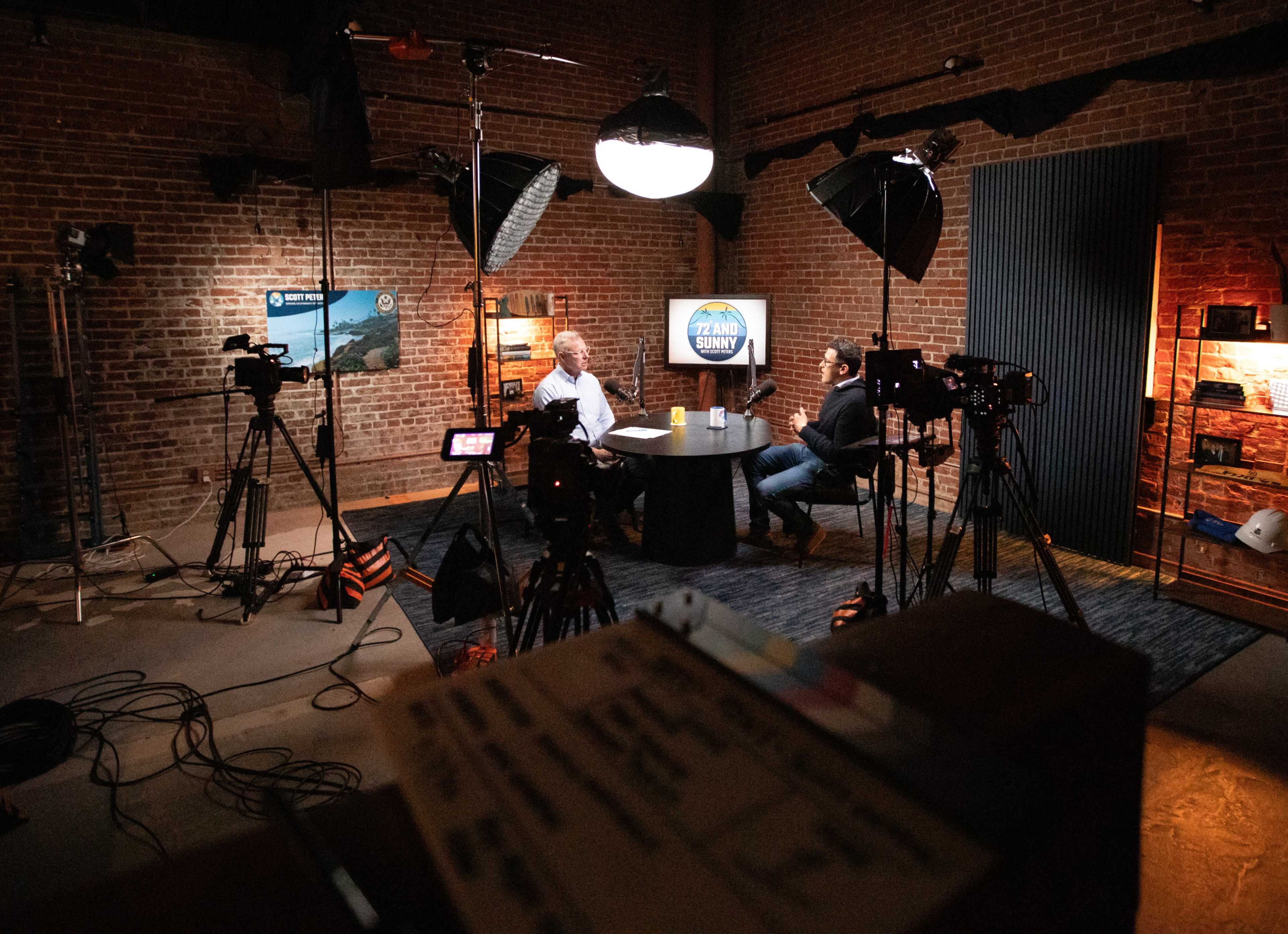 The image shows a well-equipped studio with brick walls, featuring two people seated at a table, surrounded by multiple cameras and lights.