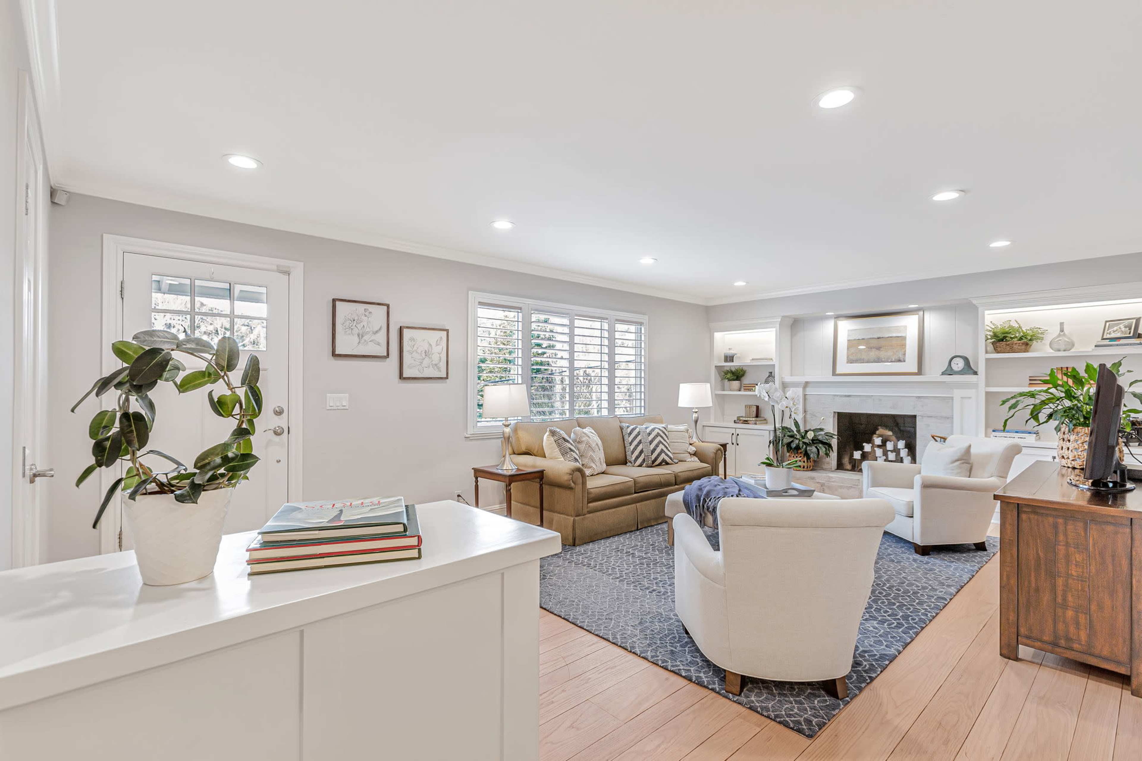 A cozy living room features a beige sofa, two white chairs, a fireplace, and natural light from large windows.