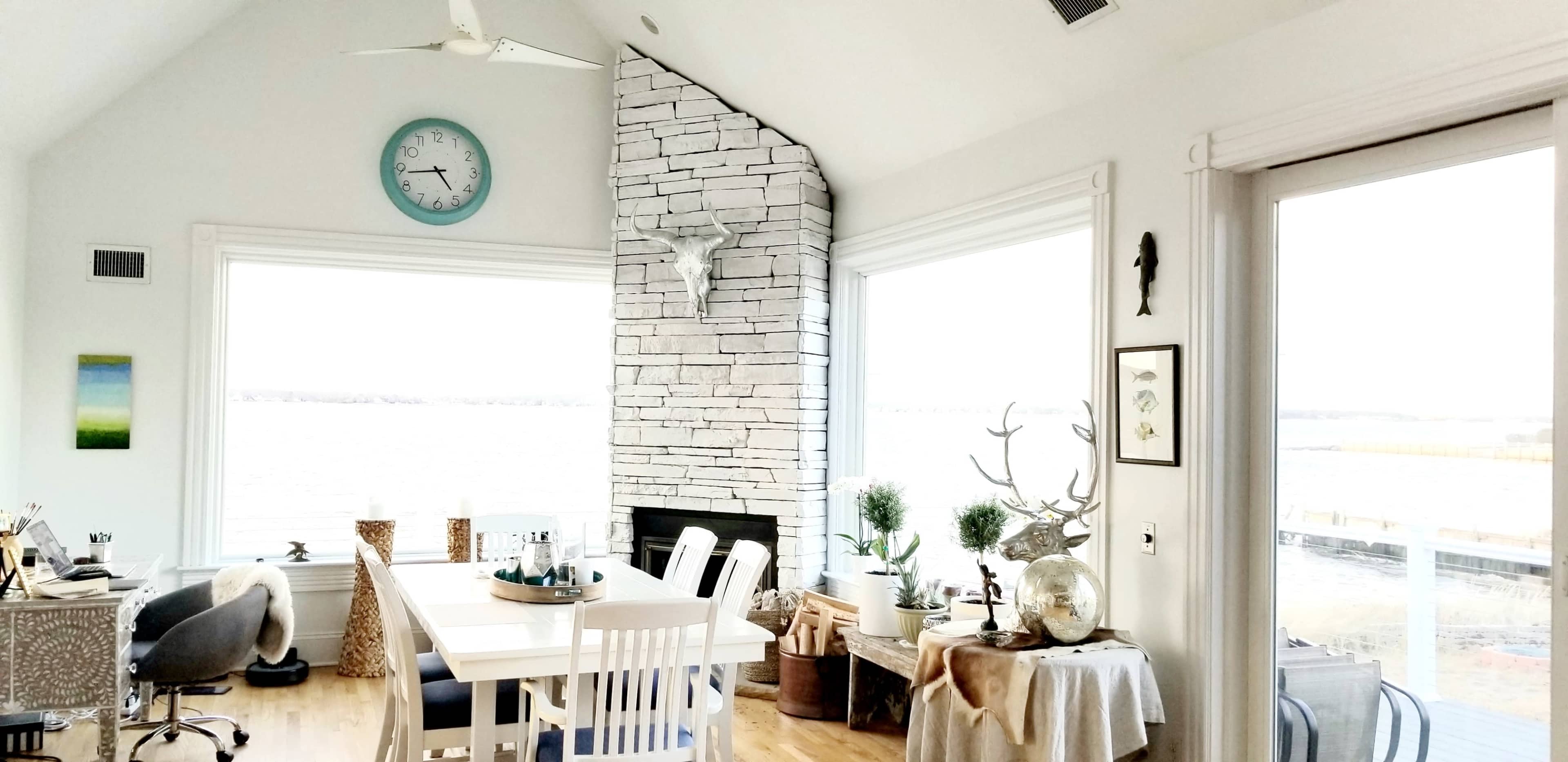A bright, airy living space with a large dining table, a stacked stone fireplace, and large windows overlooking a body of water.