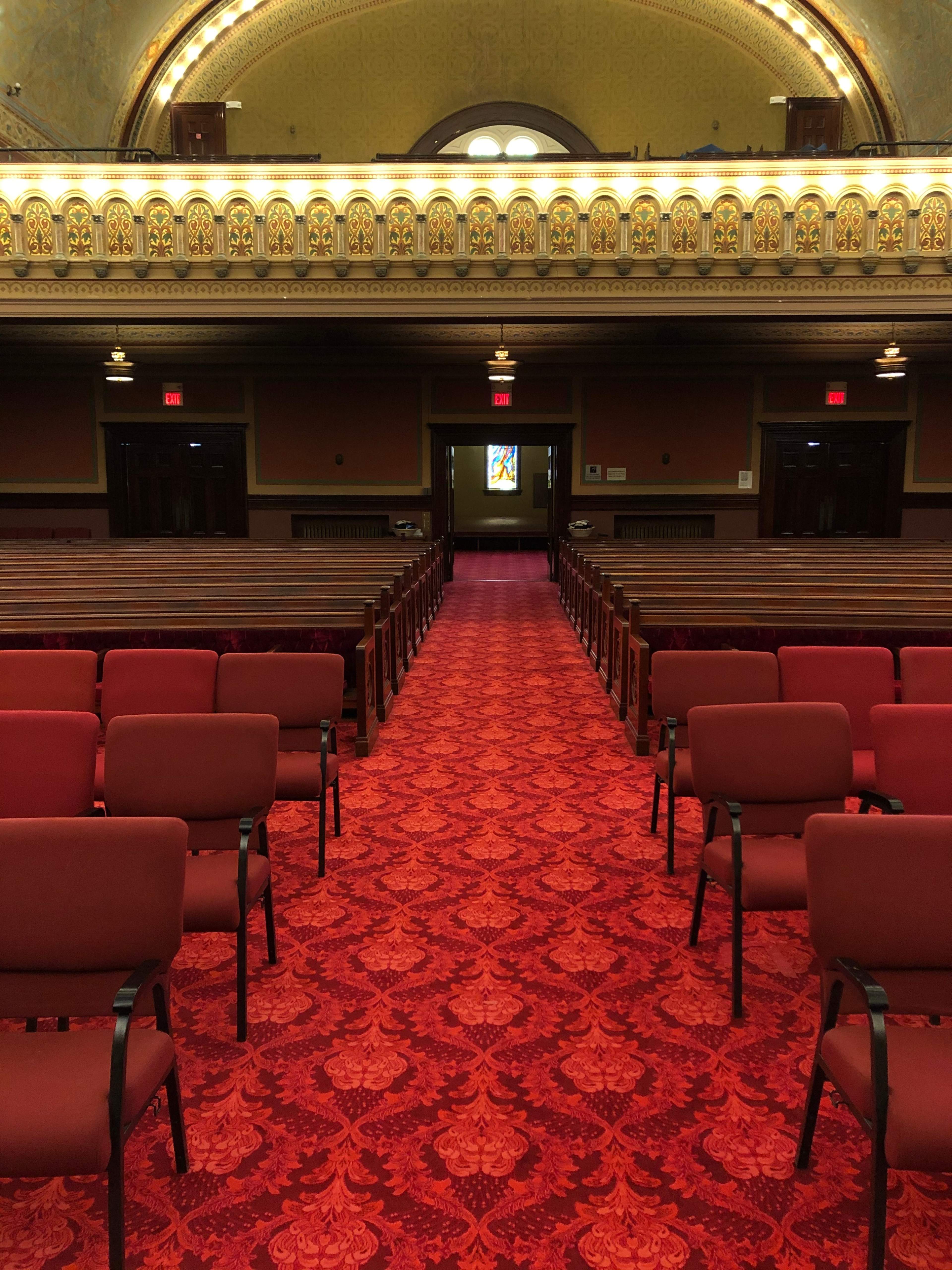 A row of empty red chairs facing a long carpeted aisle leading to a doorway with a colorful stained glass window at the end.
