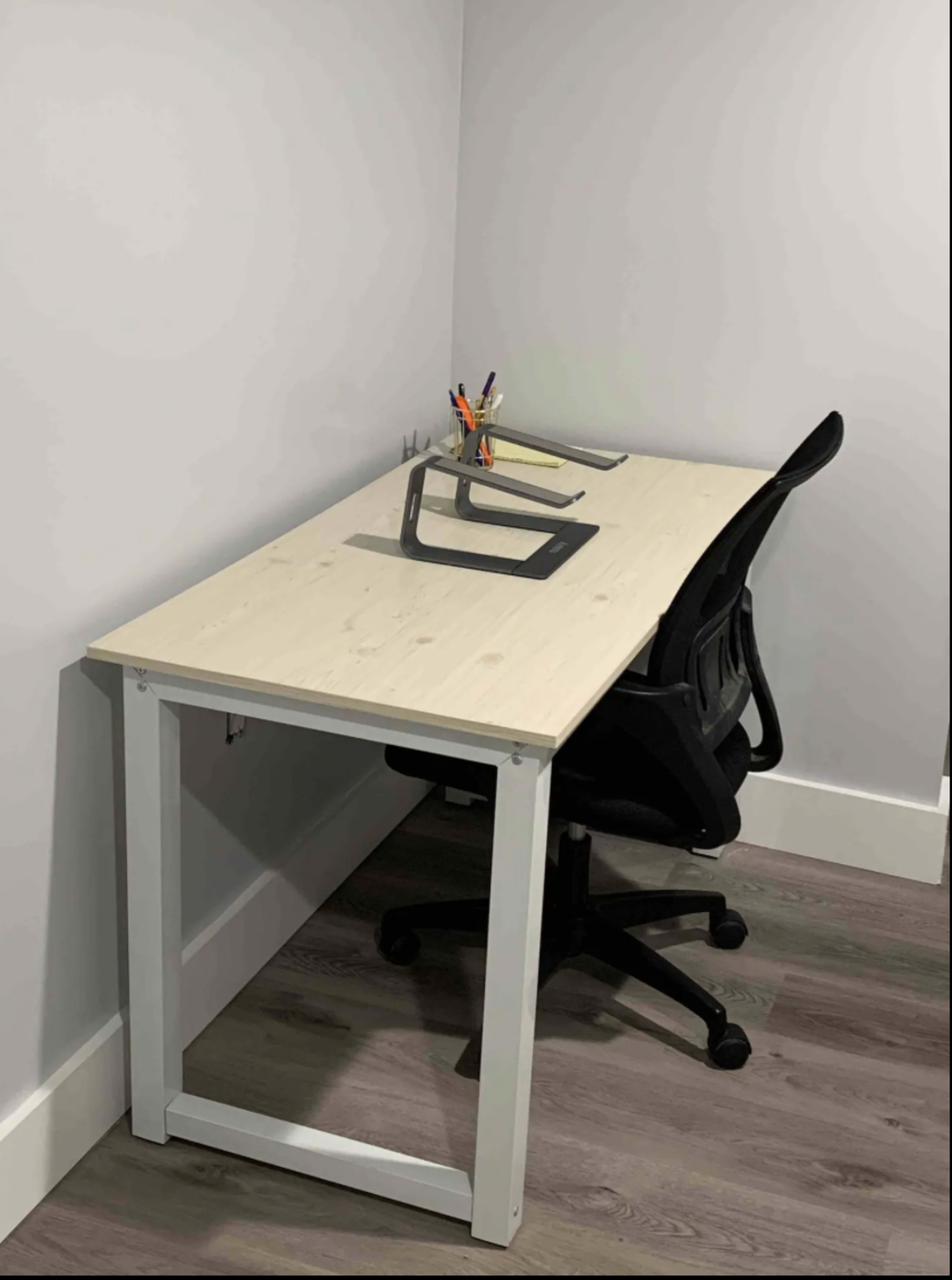 A minimalistic desk in a corner with a black office chair and a pen holder on the surface.