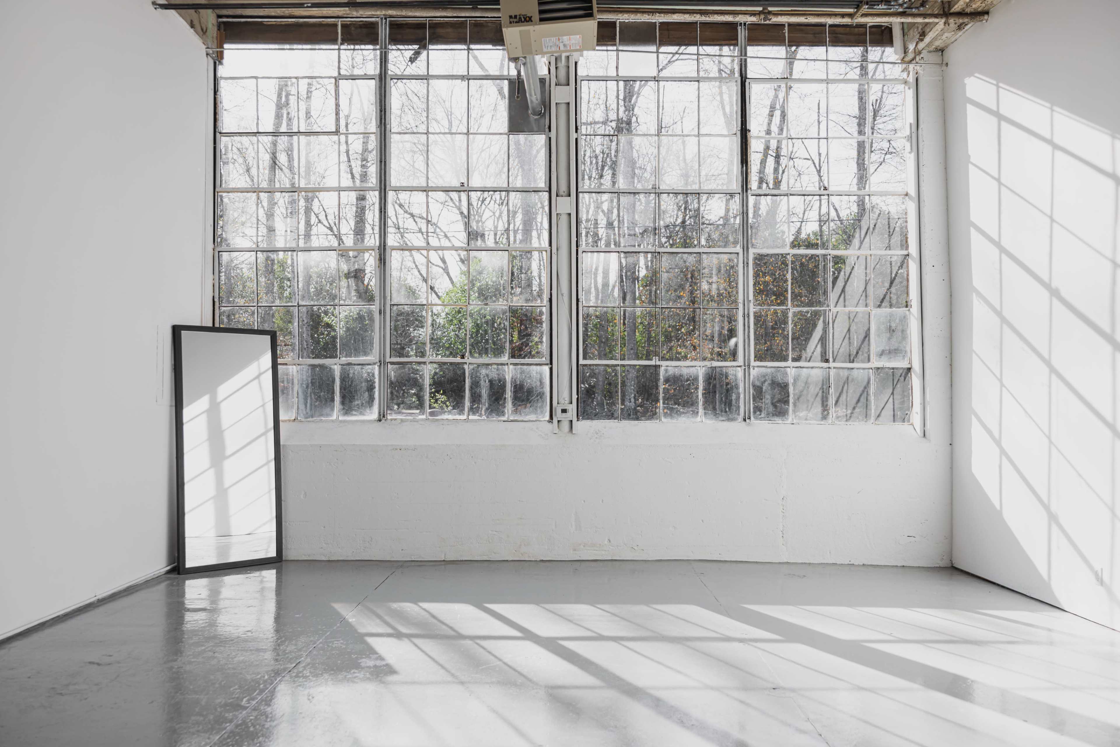 The image shows a bright, empty room with large windows and a standing mirror leaning against the wall.