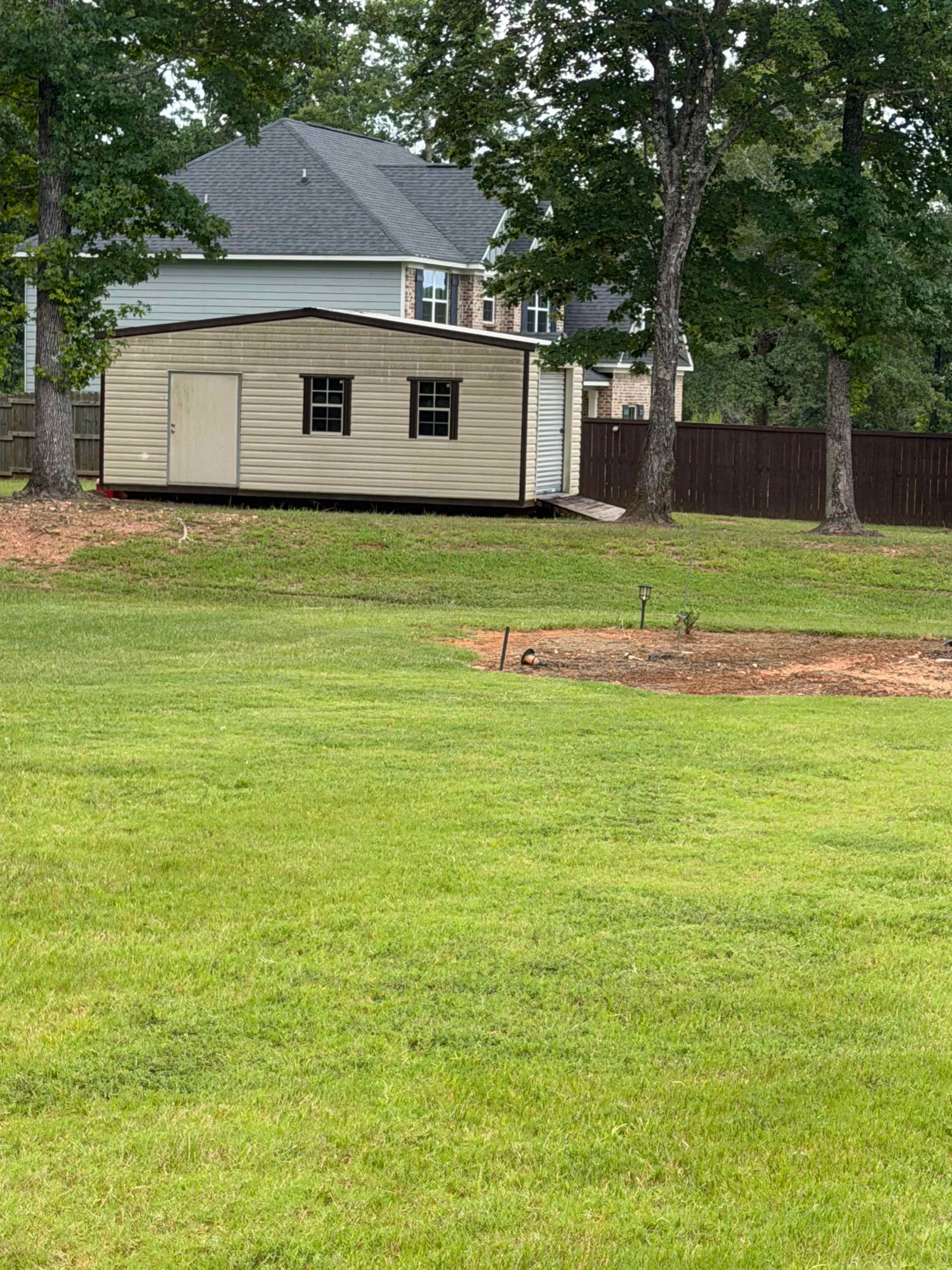 The image shows a wooden shed in a grassy yard with trees and a house in the background.