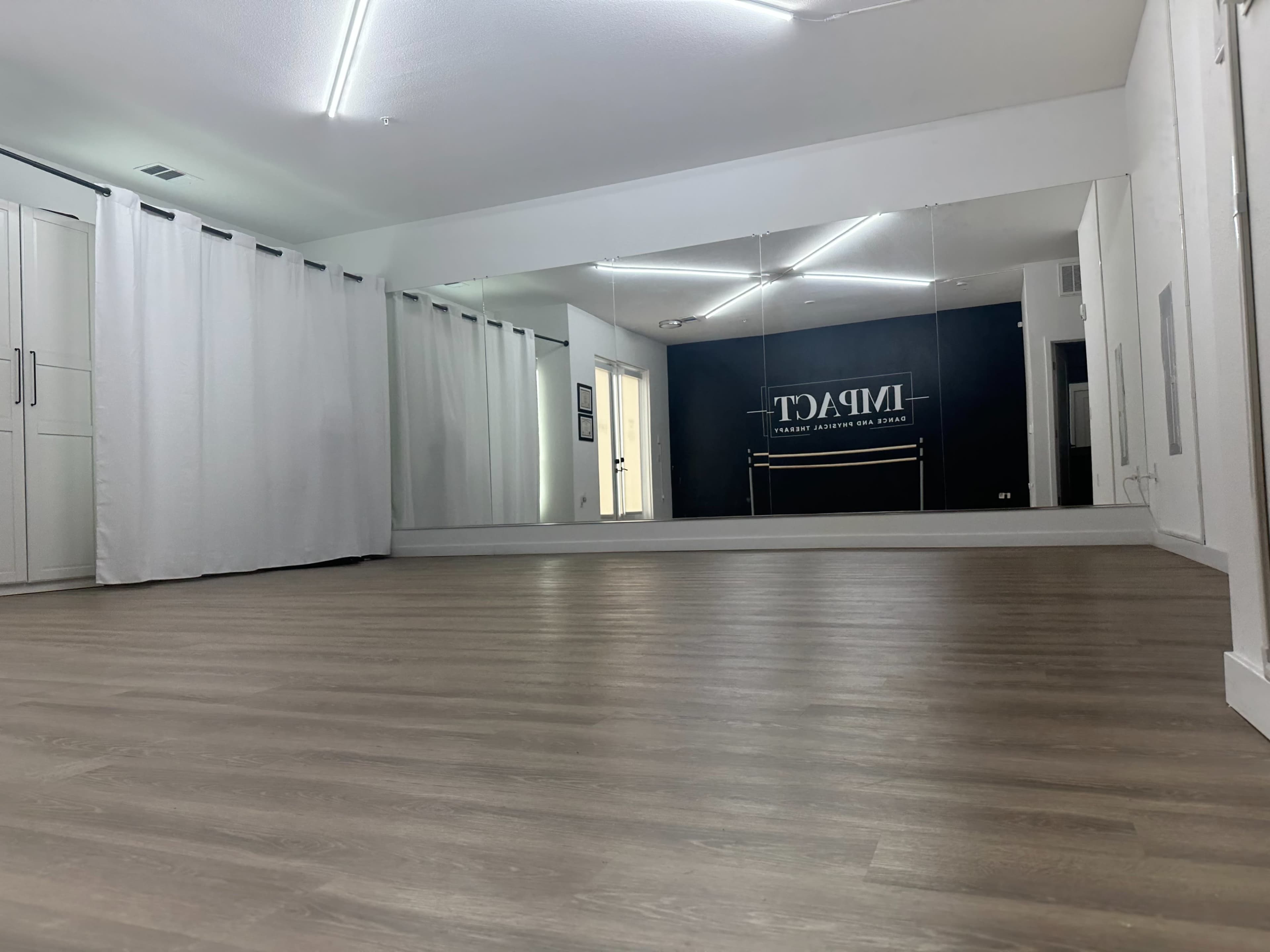 A spacious studio with wooden flooring, a large mirror on one wall, and white curtains beside cabinets.