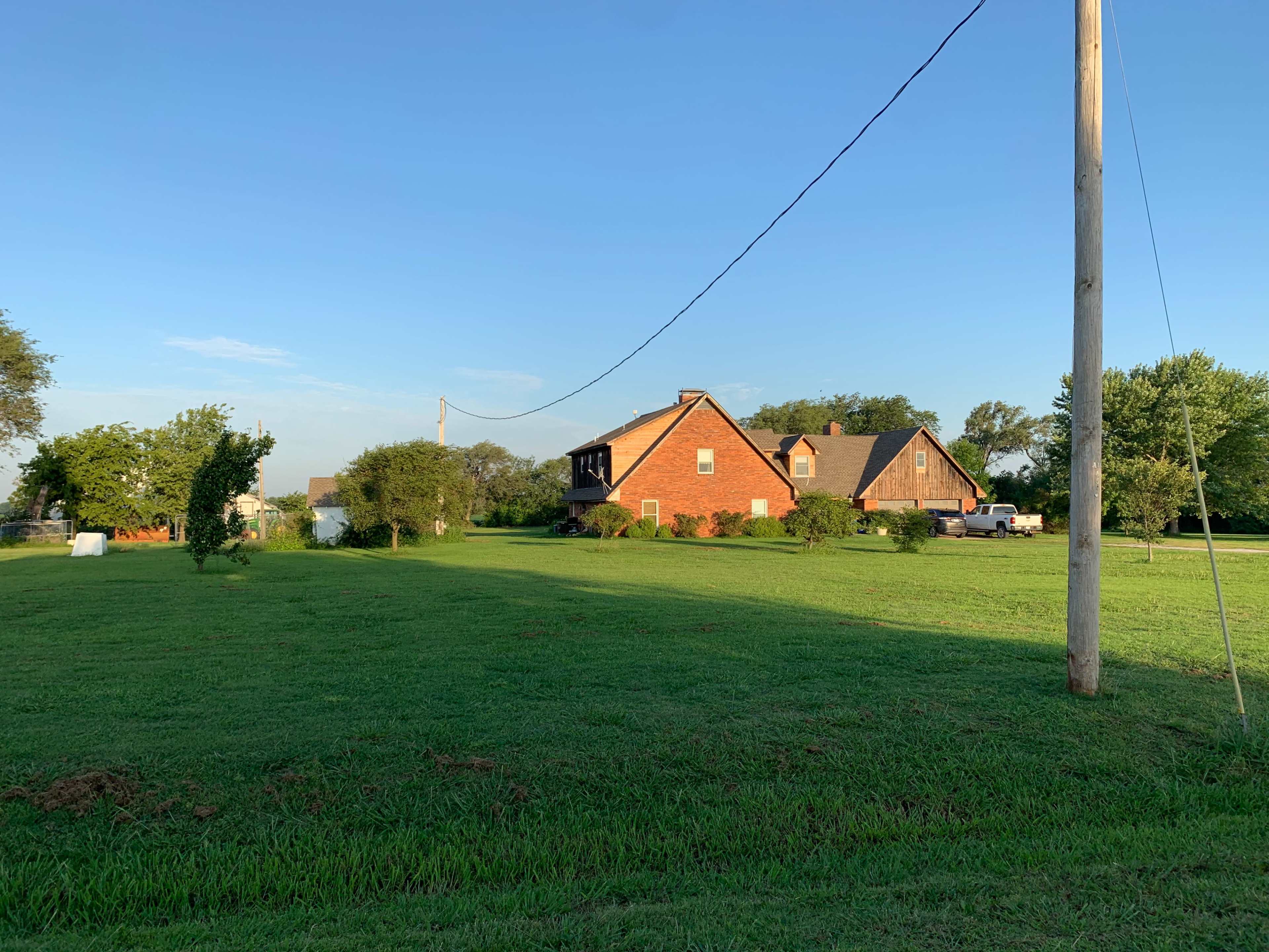 A red brick house with a sloped roof is set on a grassy lawn, surrounded by trees and utility poles under a clear blue sky.