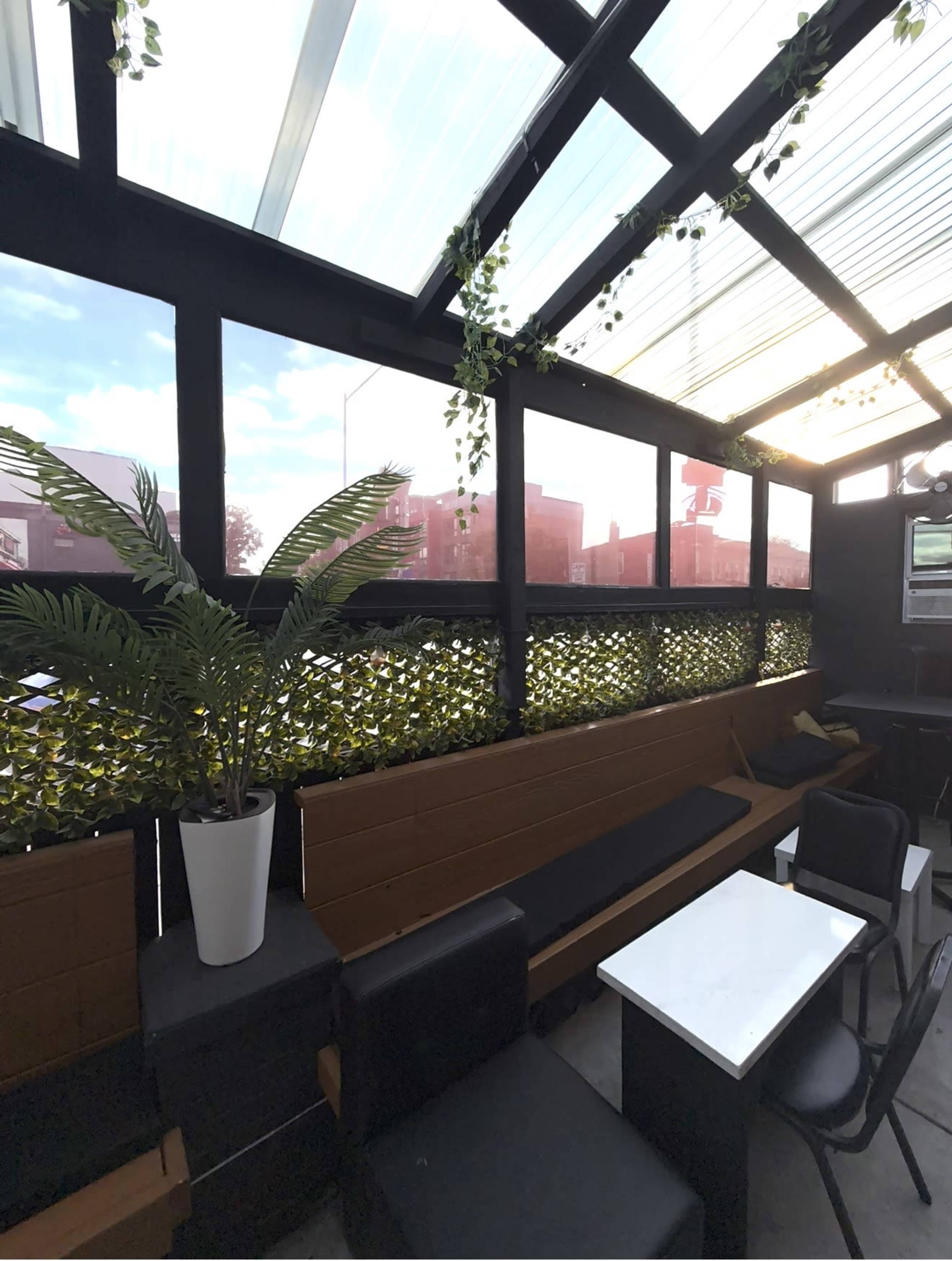 The image depicts an indoor seating area featuring a mix of wooden benches, black chairs, and a potted plant, with large windows allowing natural light to filter through.