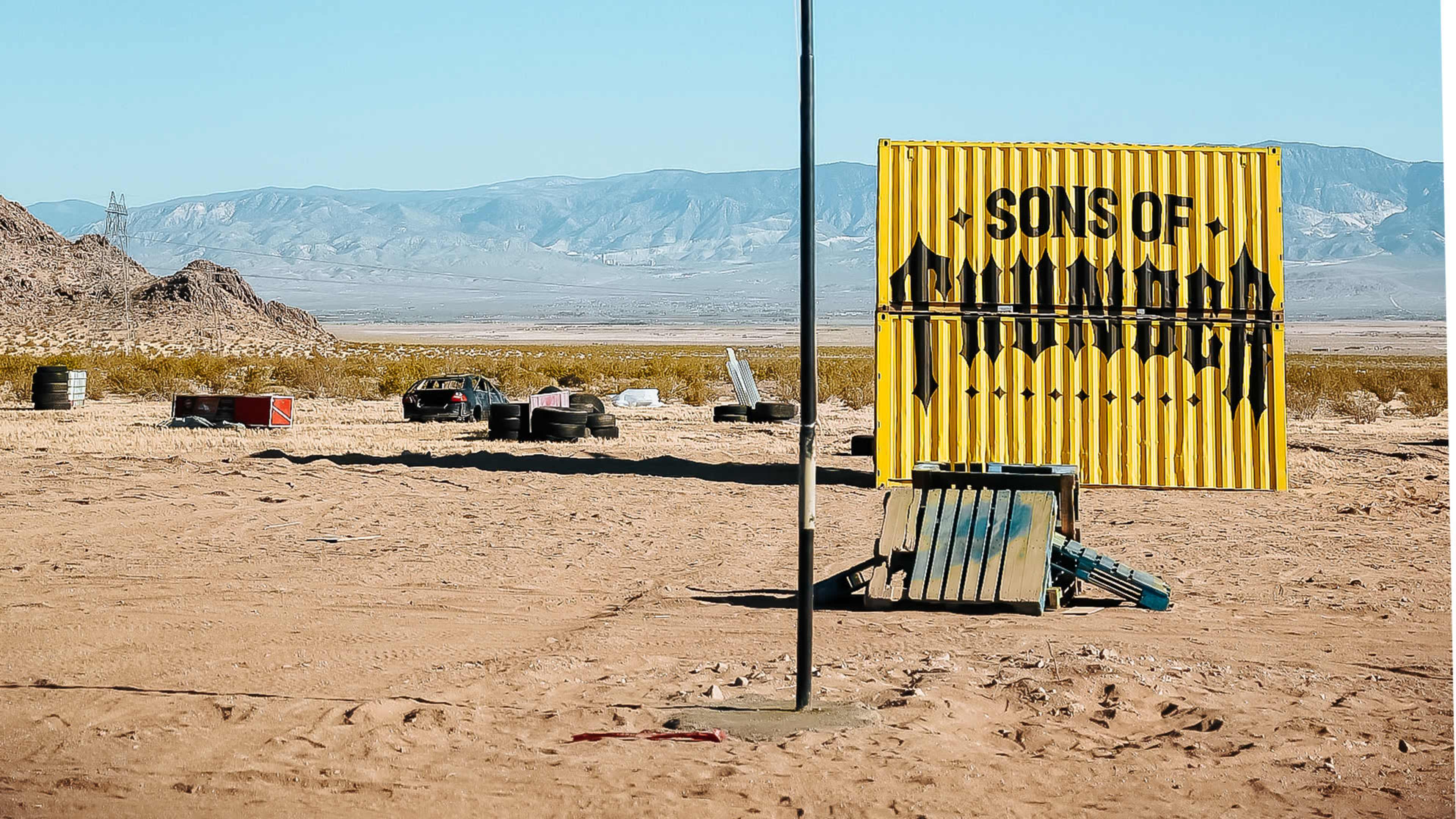 A weathered yellow sign reading "SONS OF" stands beside a dusty, abandoned landscape featuring rusted vehicles and barrels in the background.