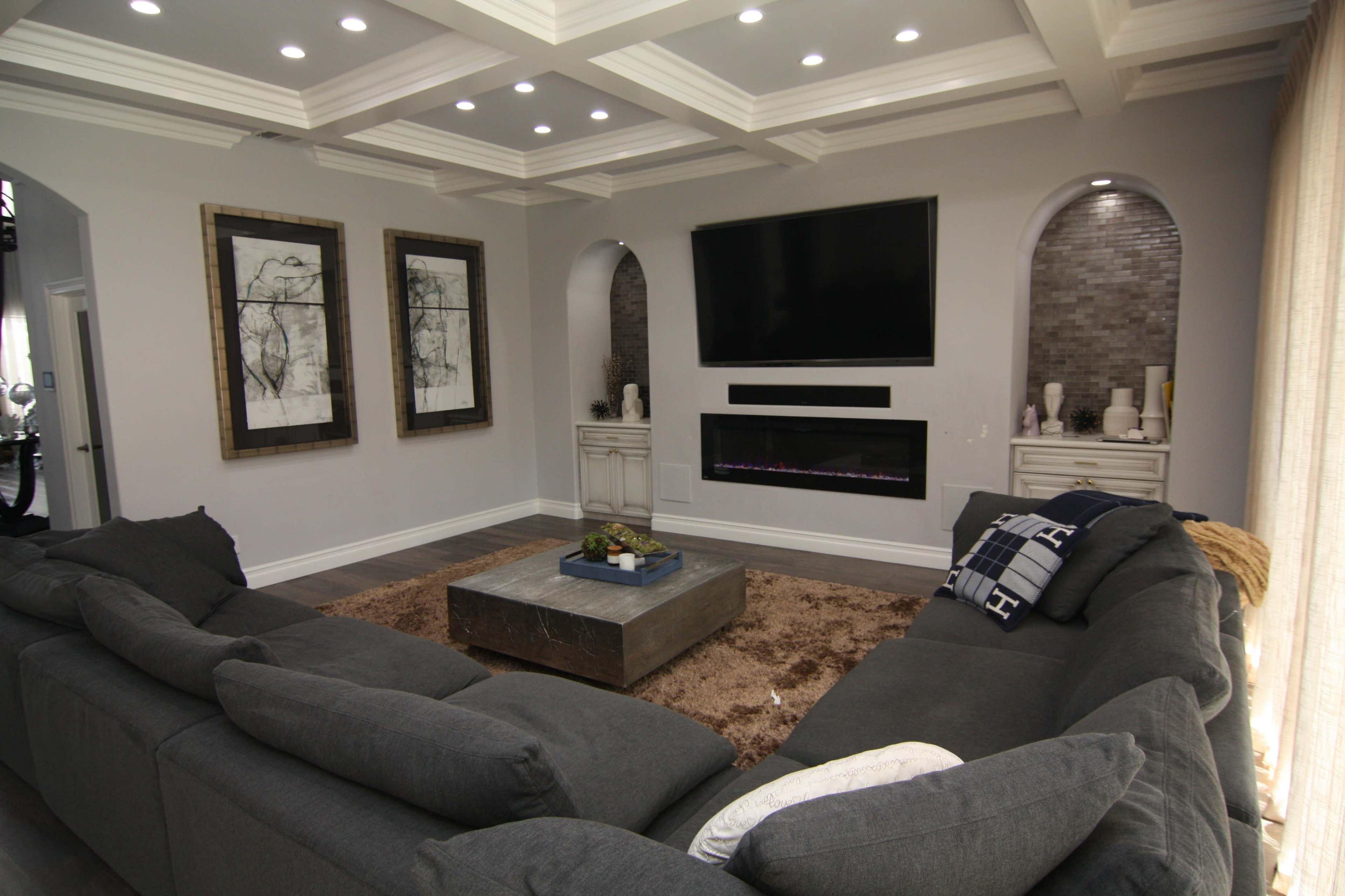 The living room features a large sectional sofa facing a mounted television above a sleek electric fireplace, with decorative artwork on the walls and a coffee table in the center.