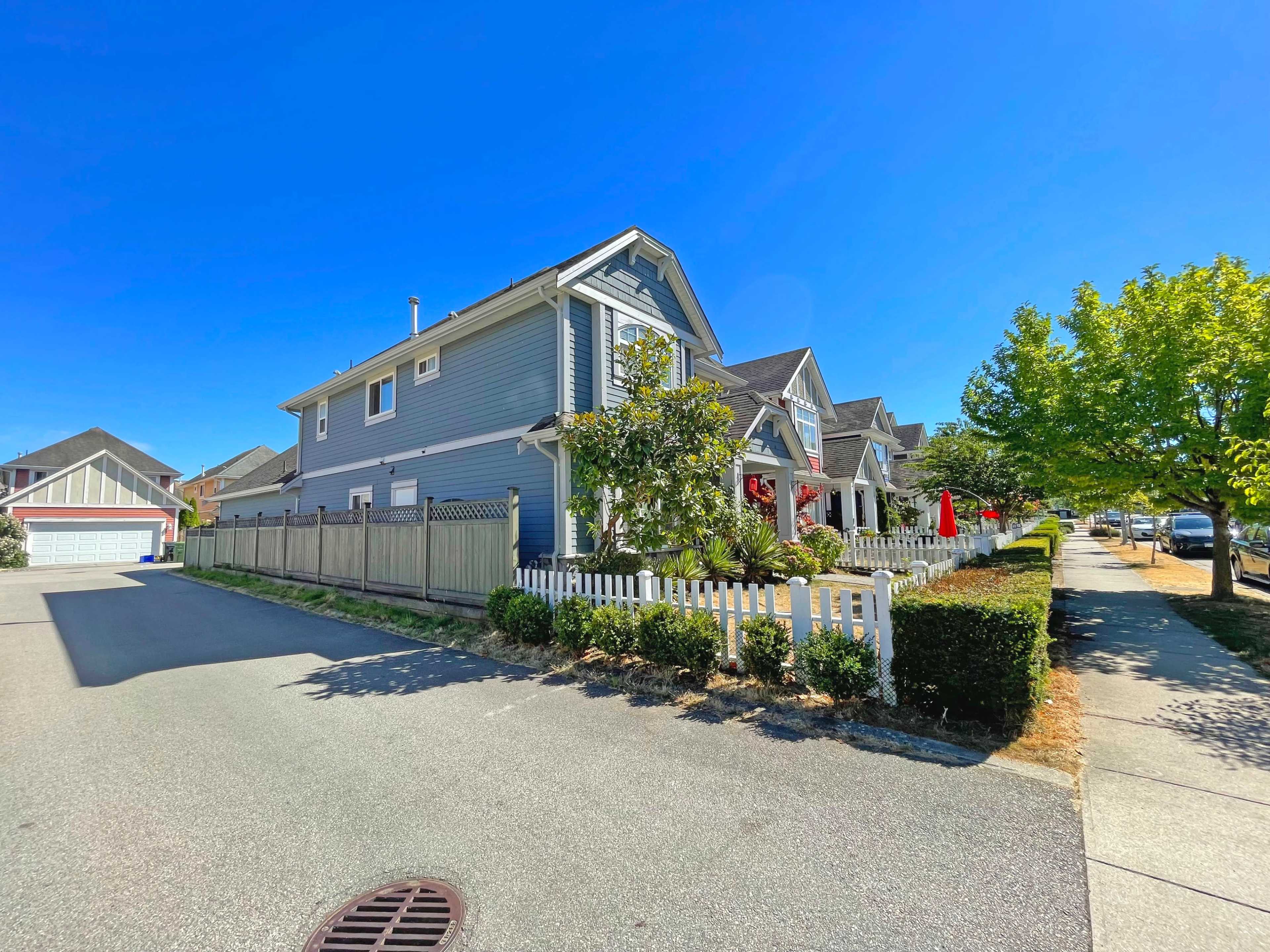 A blue two-story house with a white picket fence is situated at the corner of a street, surrounded by neatly trimmed hedges and sidewalks.