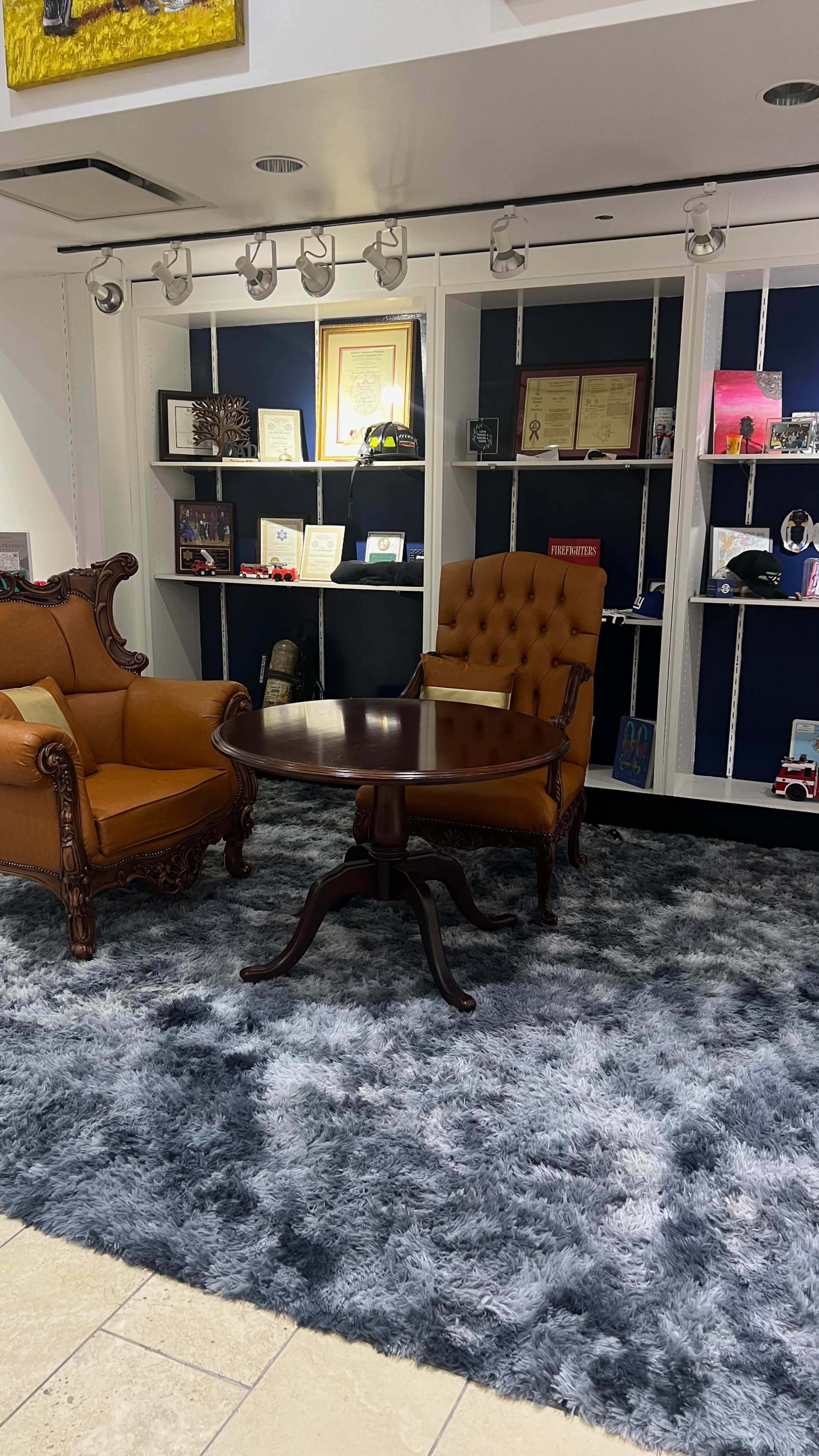 The image shows a room featuring two vintage armchairs, a round wooden table, and shelves displaying various framed items and decorations against a dark blue backdrop.