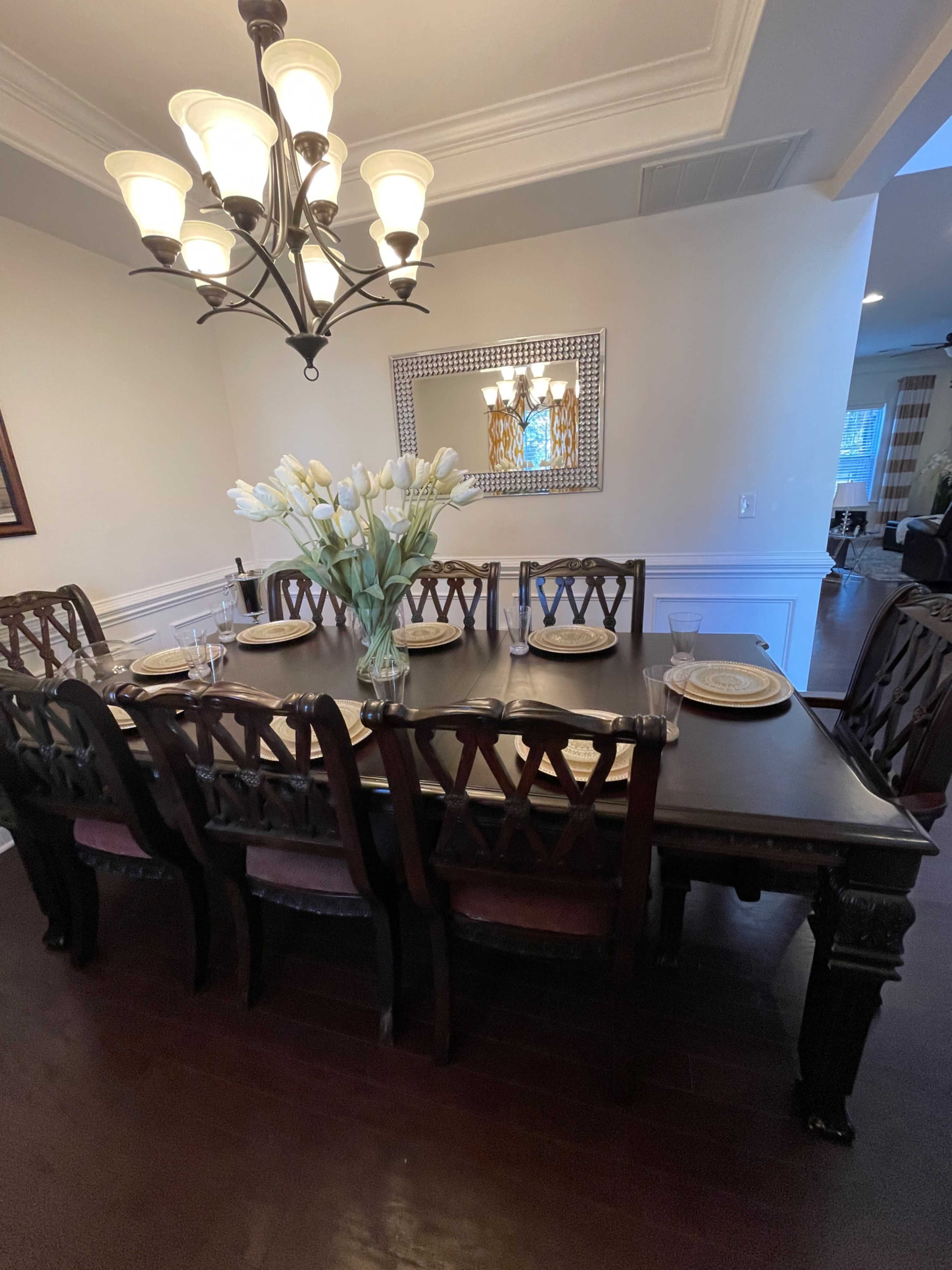 A dark wooden dining table is set with plates and silverware, surrounded by eight chairs, under a chandelier with multiple light bulbs.