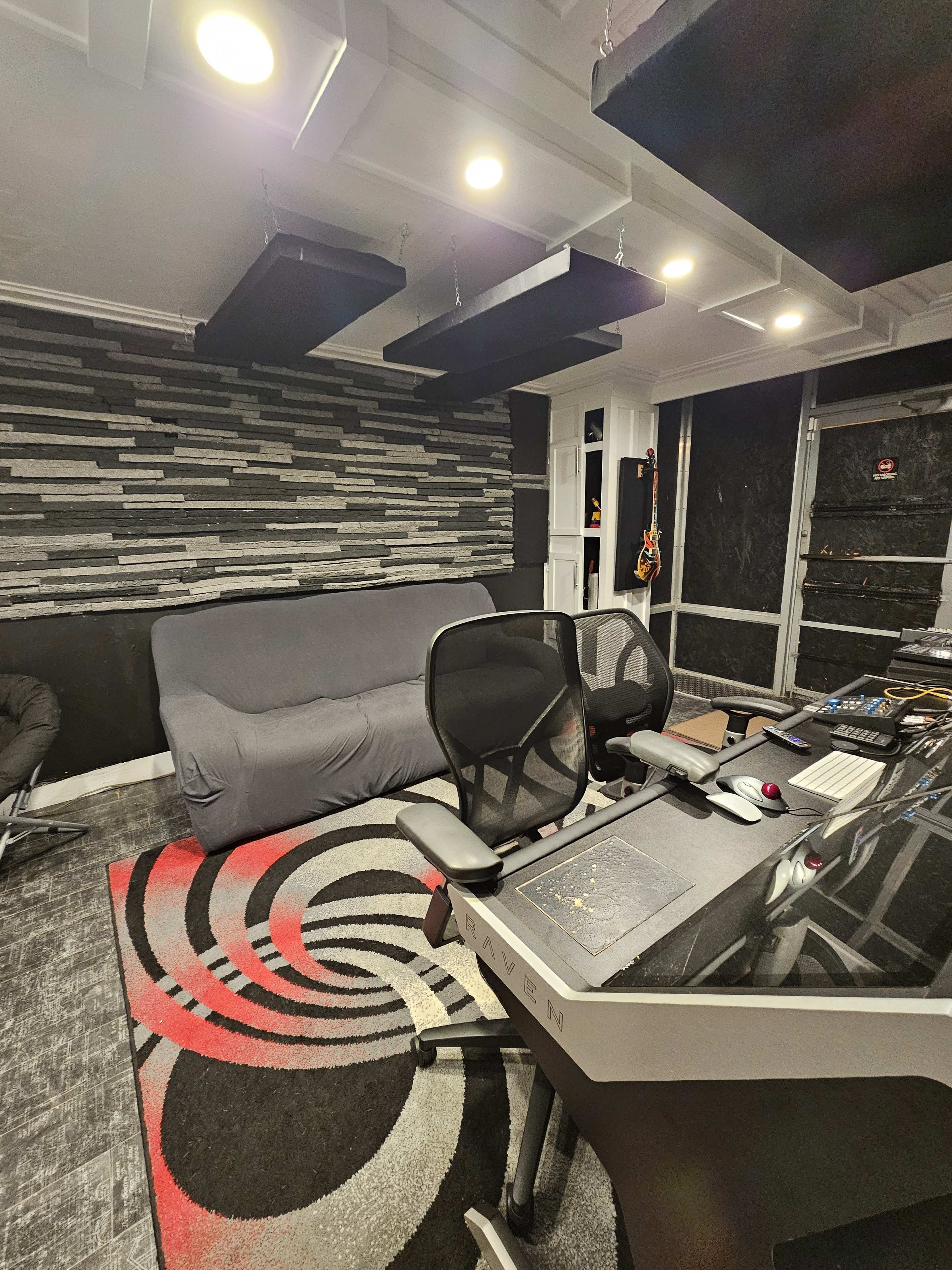 The image shows a recording studio with a dark color scheme, featuring a black and gray patterned wall, a couch, and a desk with audio equipment and two office chairs.