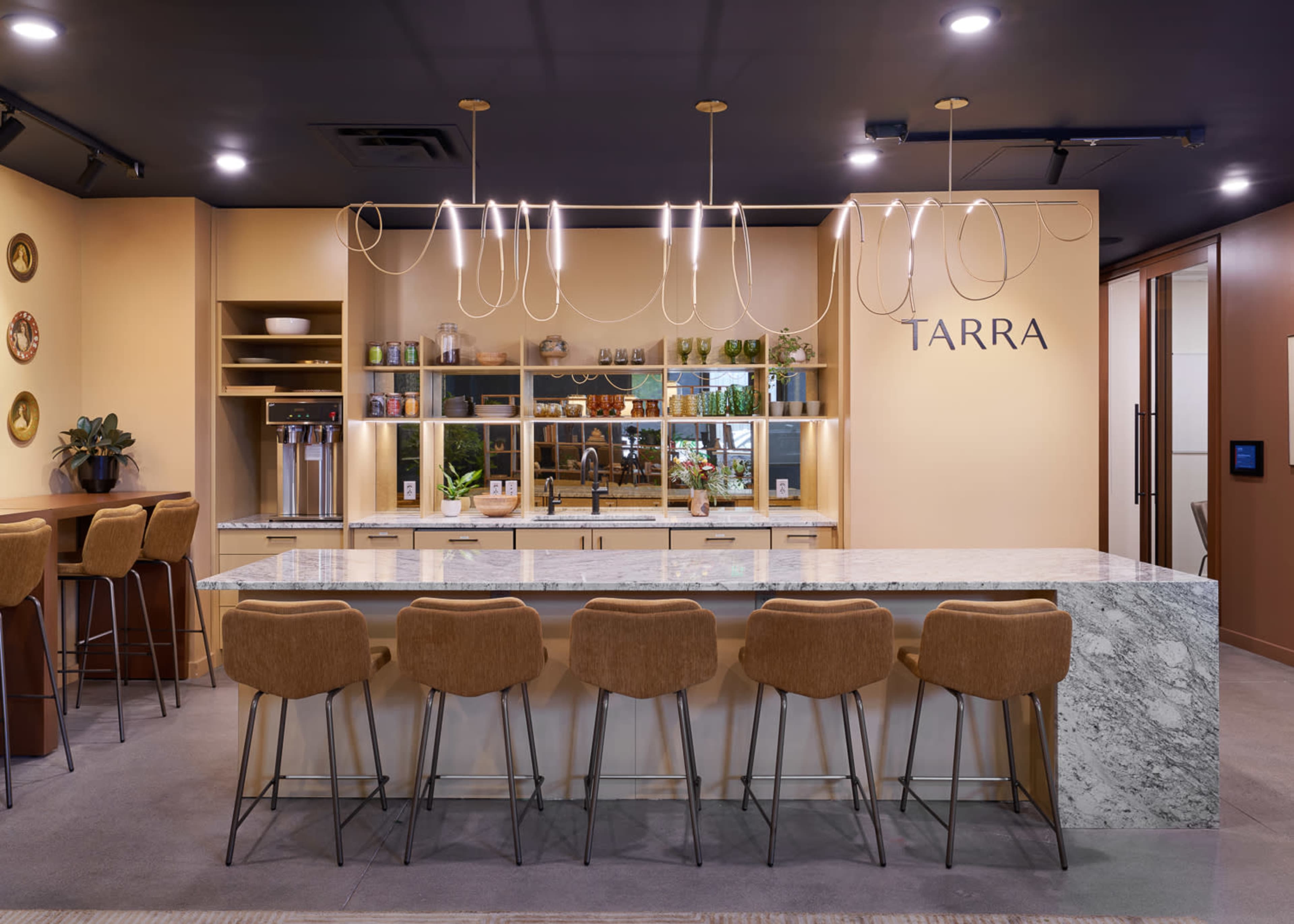 A modern kitchen area with a long marble-topped bar, stylish seating, and shelving displaying various items.