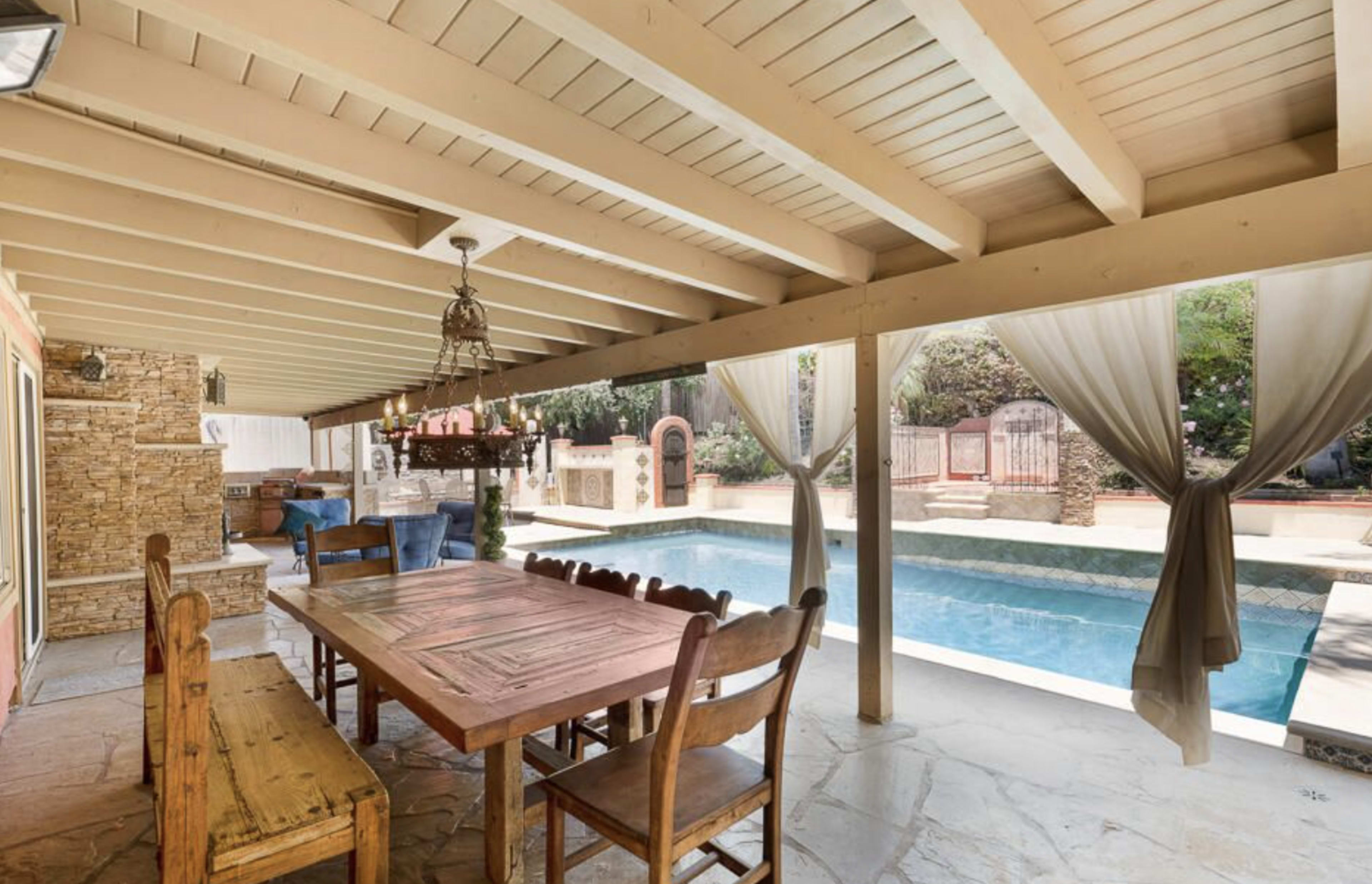 A covered patio area with a wooden dining table near a pool, flanked by stone walls and draped curtains.