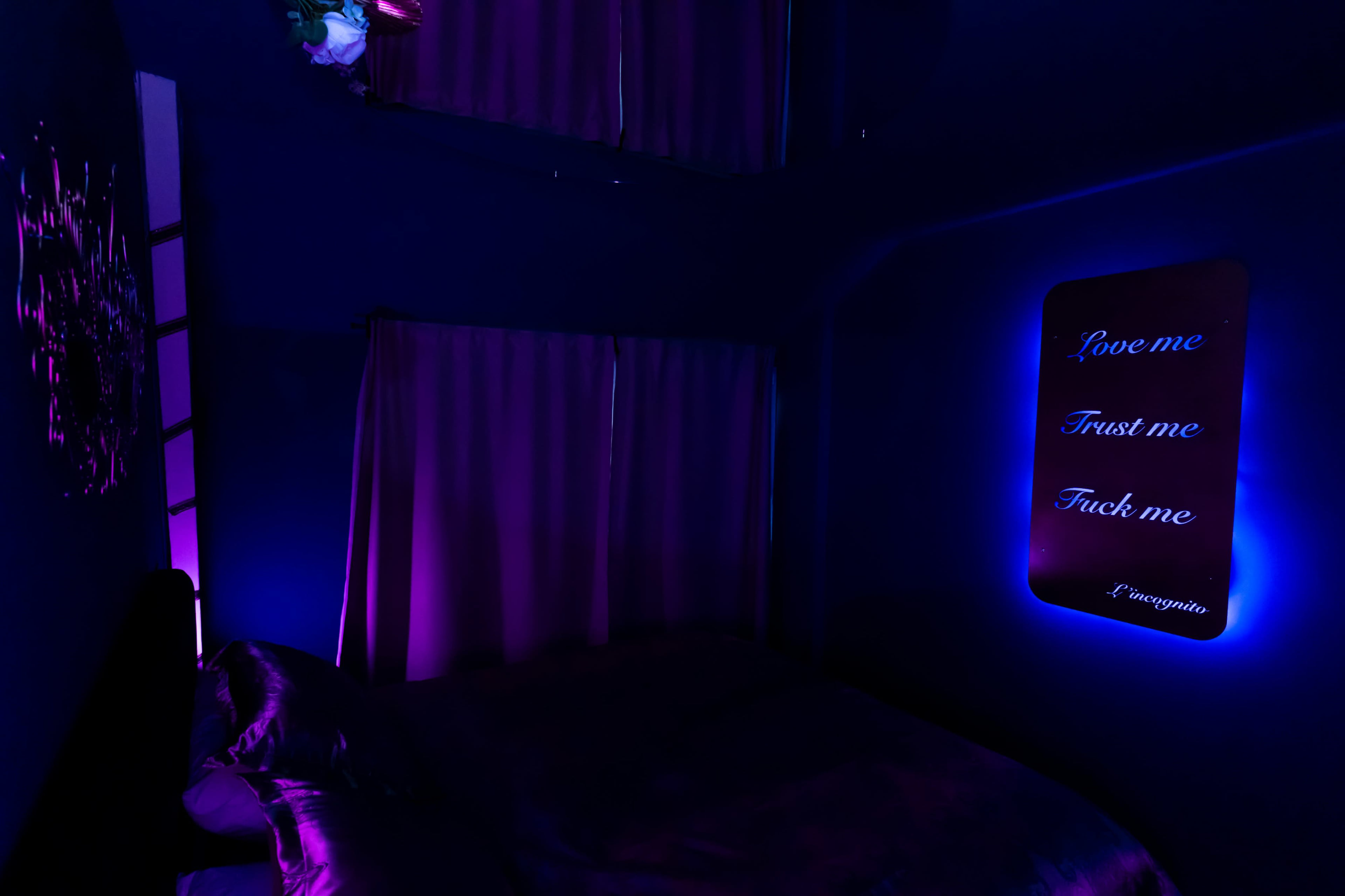 A dimly lit bedroom features purple walls, curtains, and neon text on the wall that reads "Love me, Trust me, Fuck me."