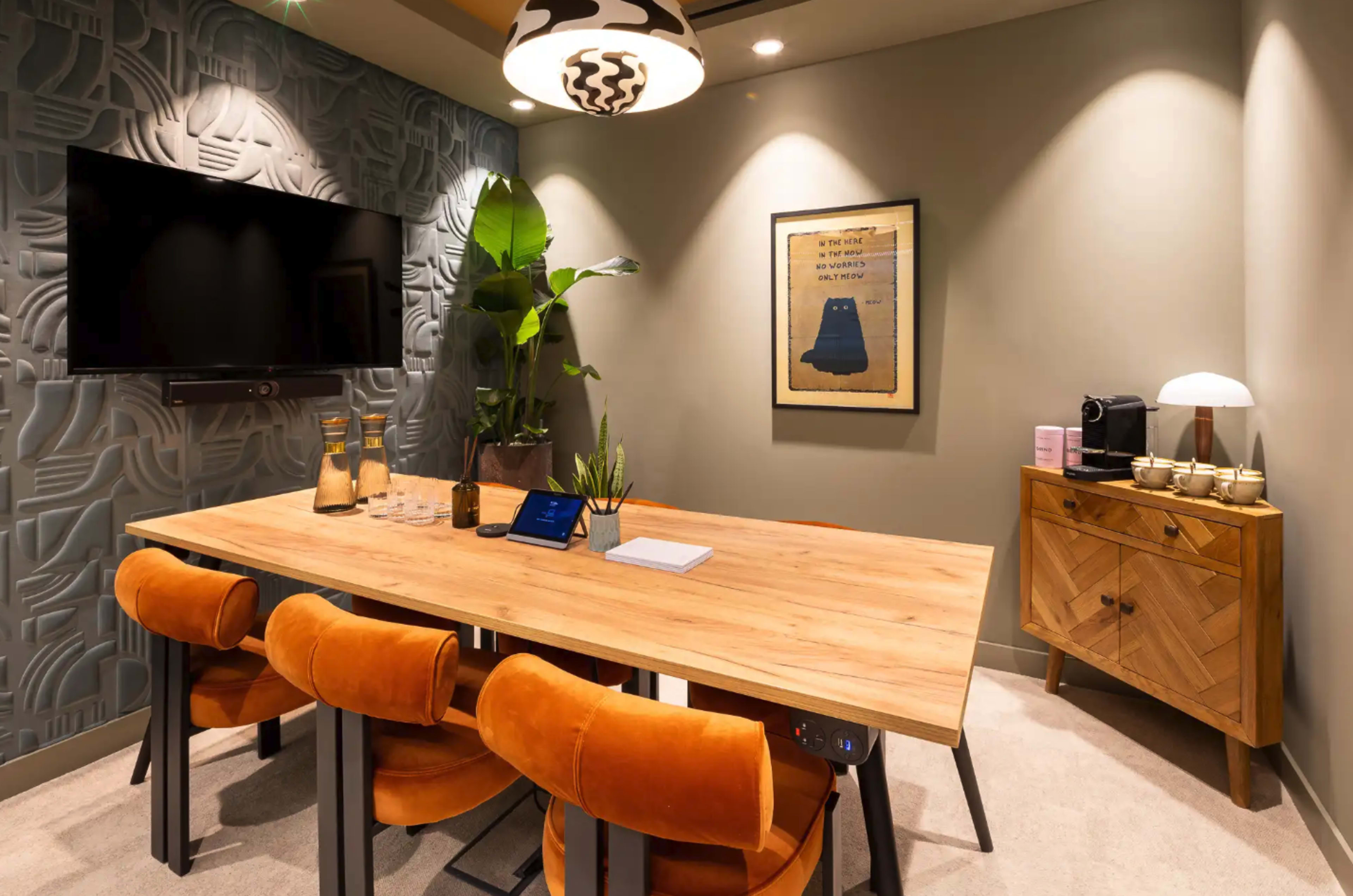 The image shows a small meeting room featuring a wooden table surrounded by orange chairs, a television mounted on the wall, and decorative items including a plant and artwork.