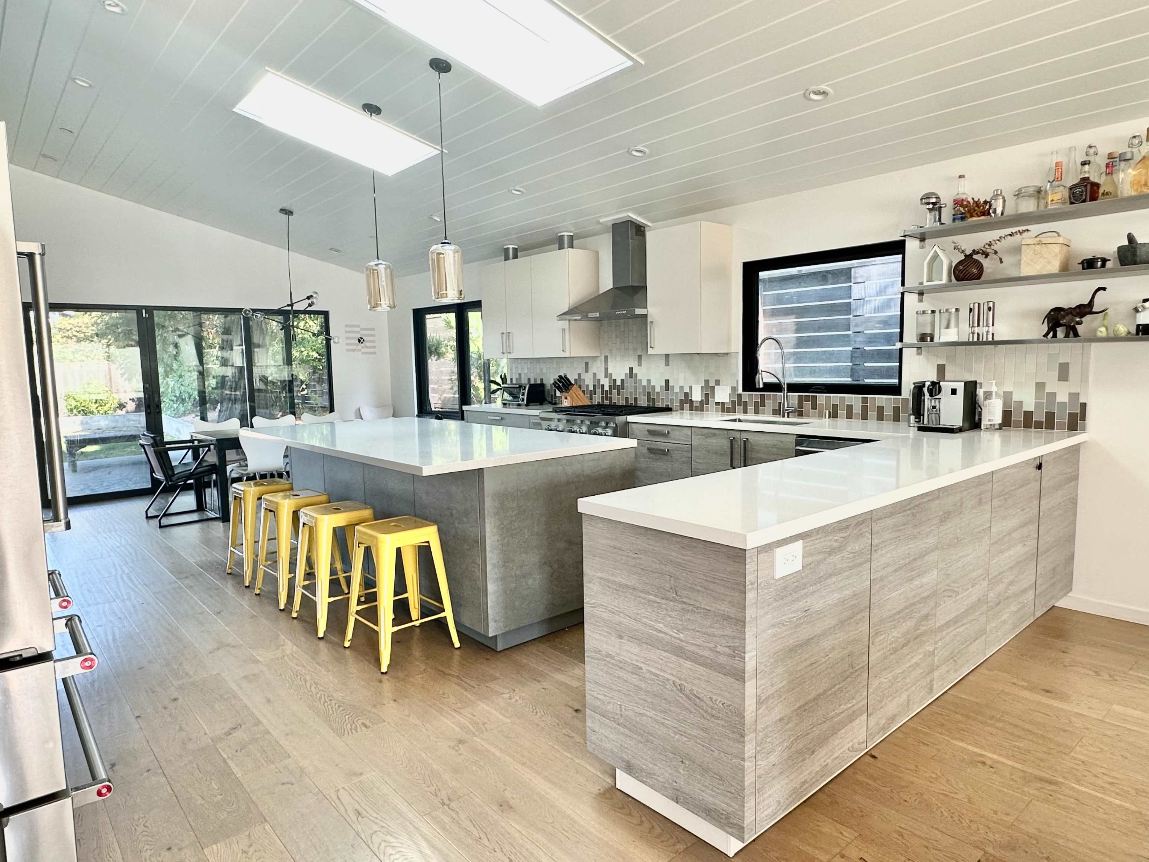 A modern kitchen features a large island with gray and white surfaces, yellow stools, and an open layout with large windows.