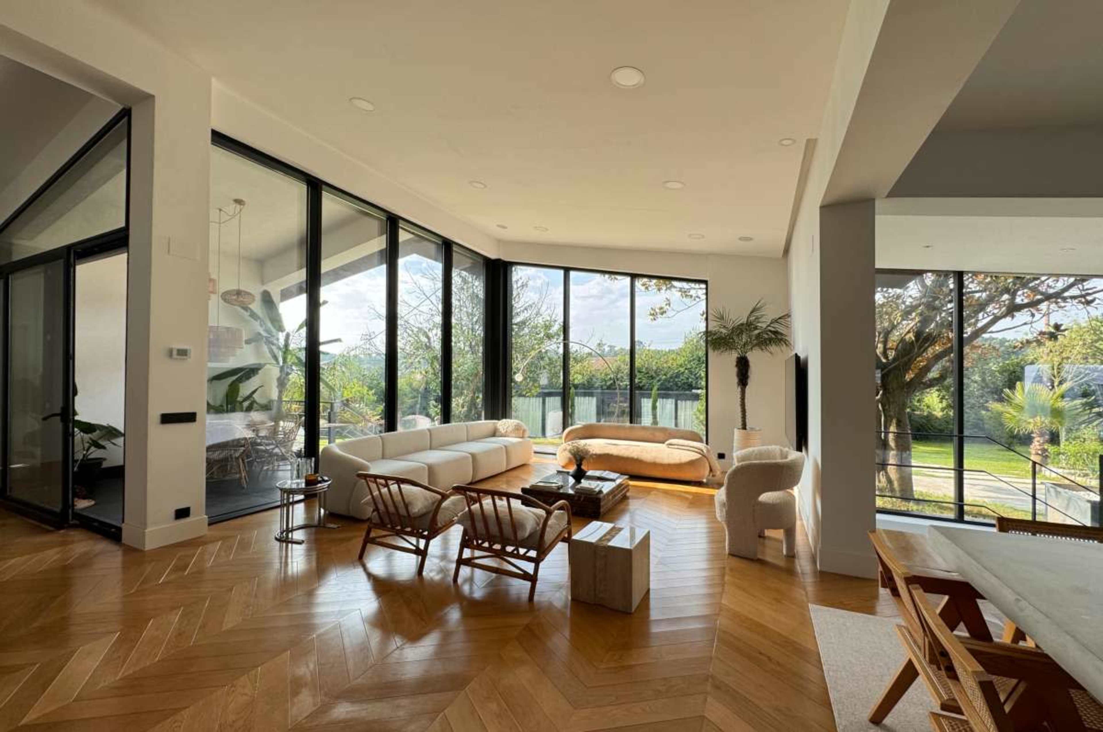 The image shows a modern living room with large glass windows, hardwood flooring, and contemporary furniture overlooking a green outdoor space.