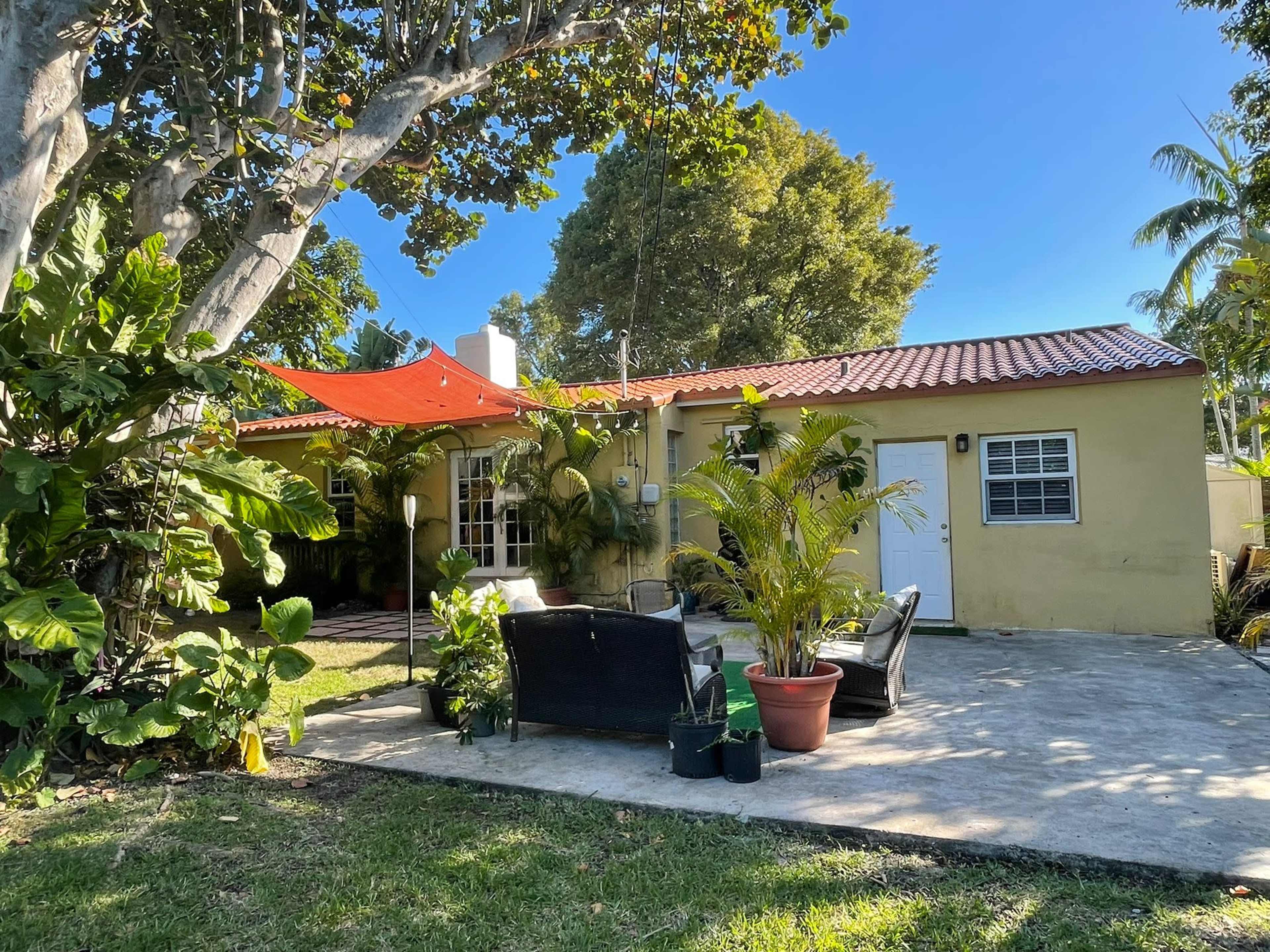 A small house with a red awning is surrounded by tropical plants and features a patio area with seating furniture.