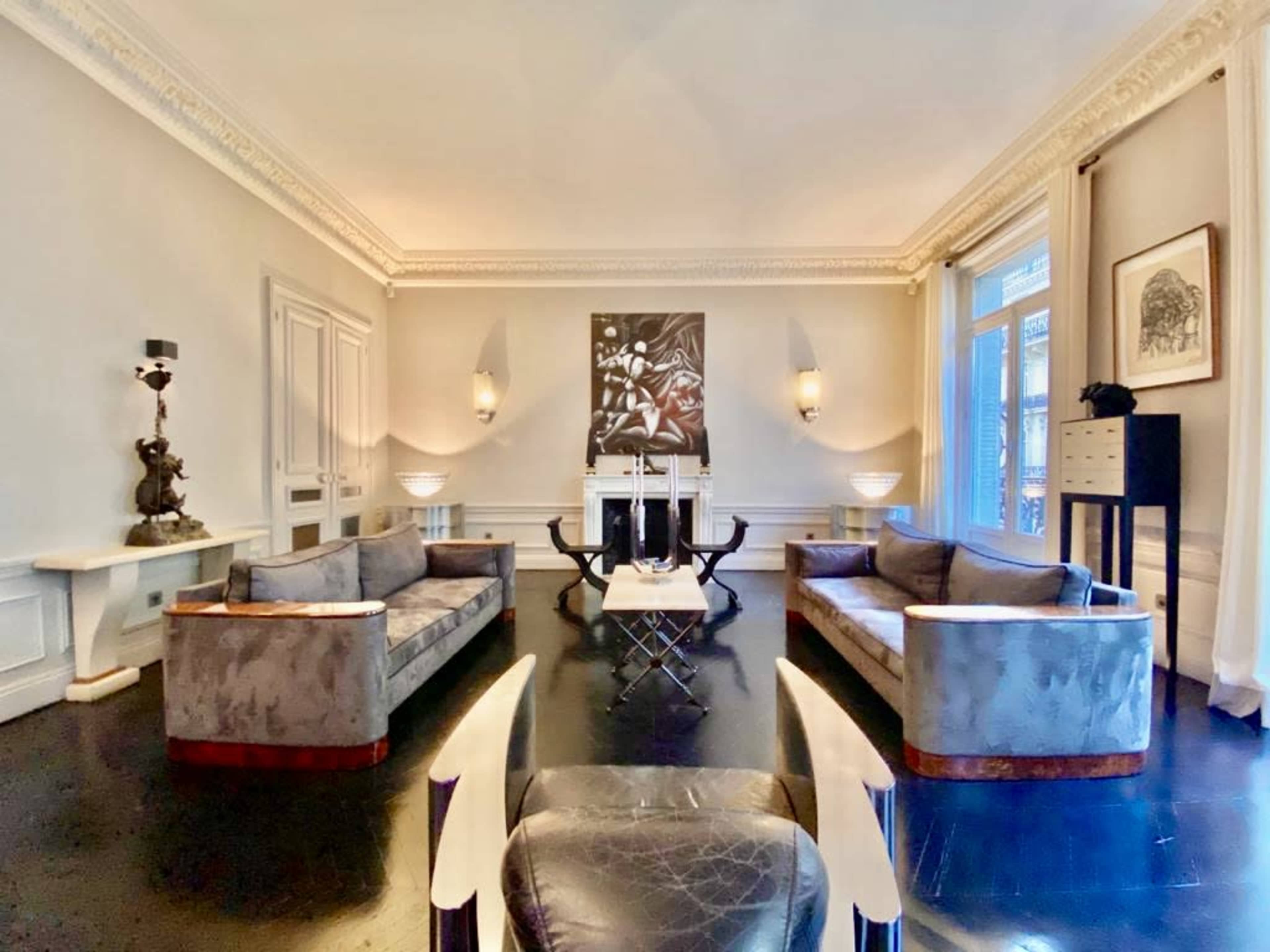 The image shows a spacious living room featuring two gray sofas, a central glass coffee table, and a large artwork on the wall.
