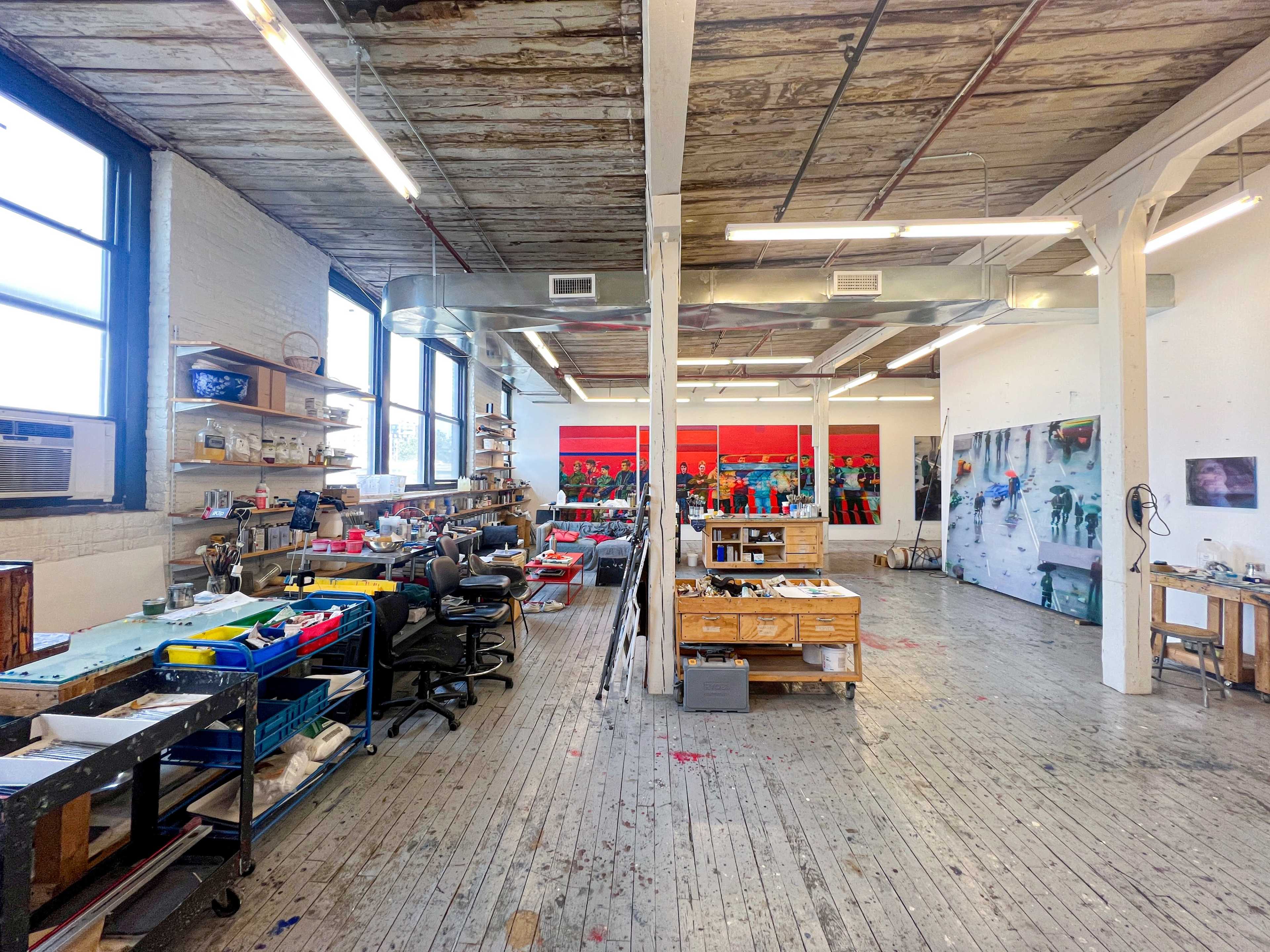 The image shows an art studio with wooden flooring, large windows, and various workstations filled with tools and materials, along with large paintings displayed on the walls.