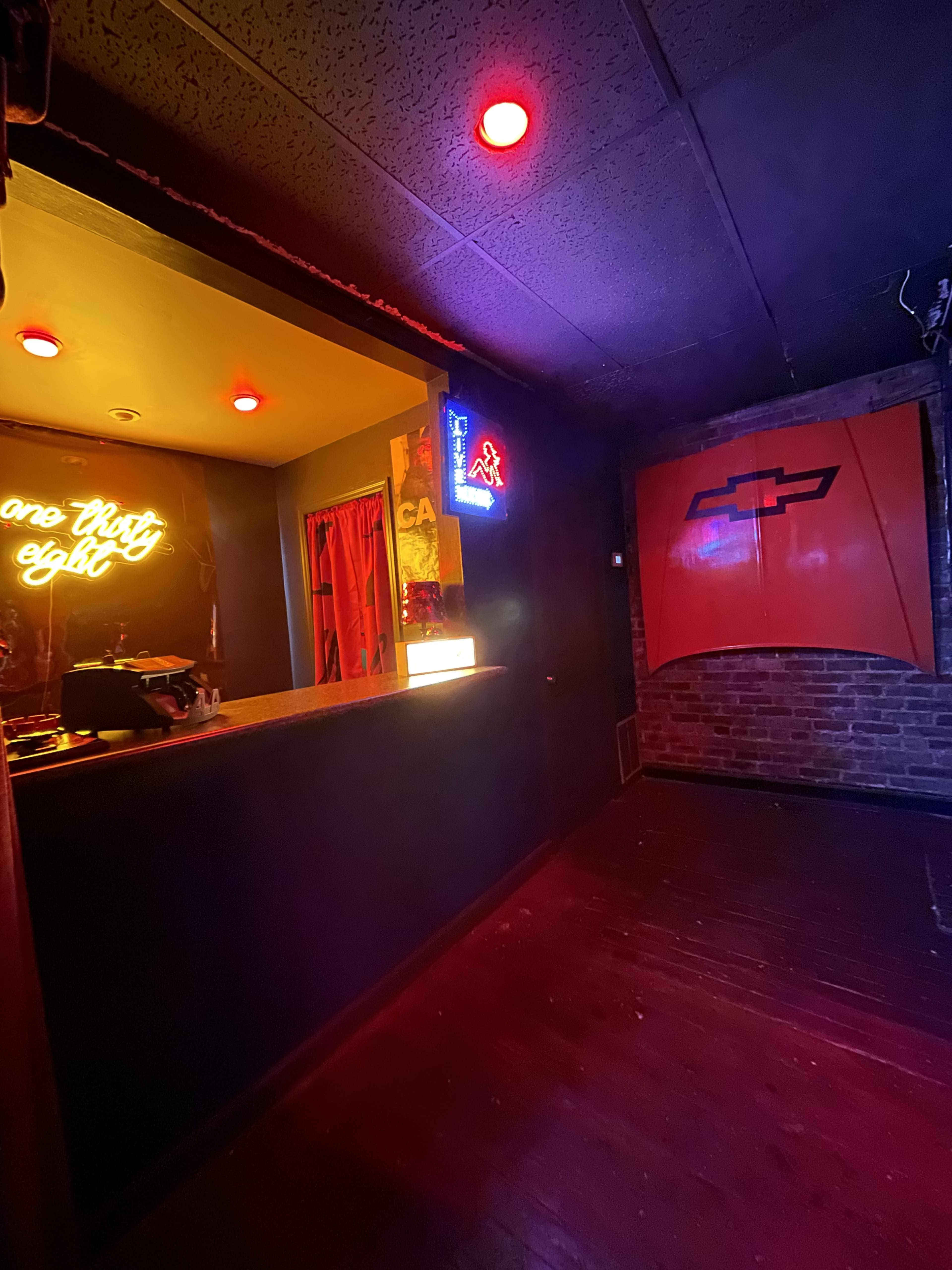 A dimly lit room with a bar area featuring neon signs, red lighting, and industrial-style brick walls.