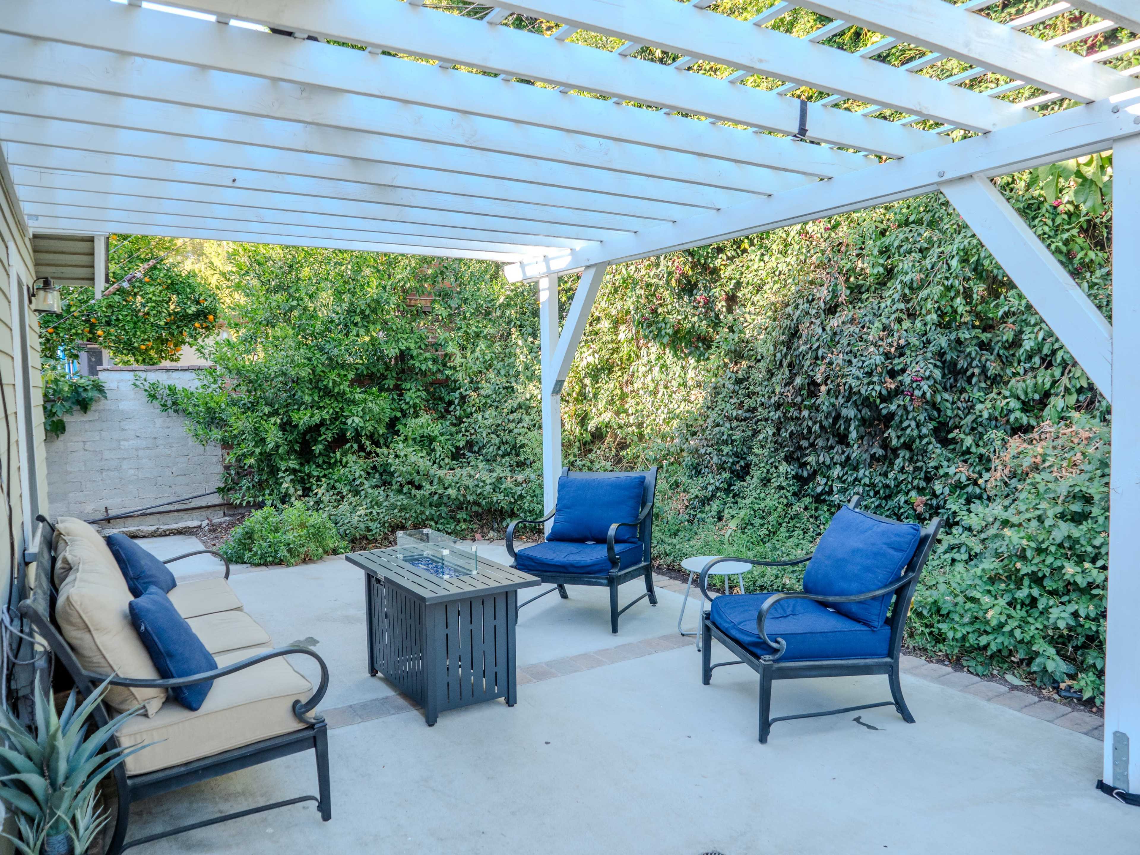 A patio area with a white pergola, outdoor seating, and a central table surrounded by greenery.
