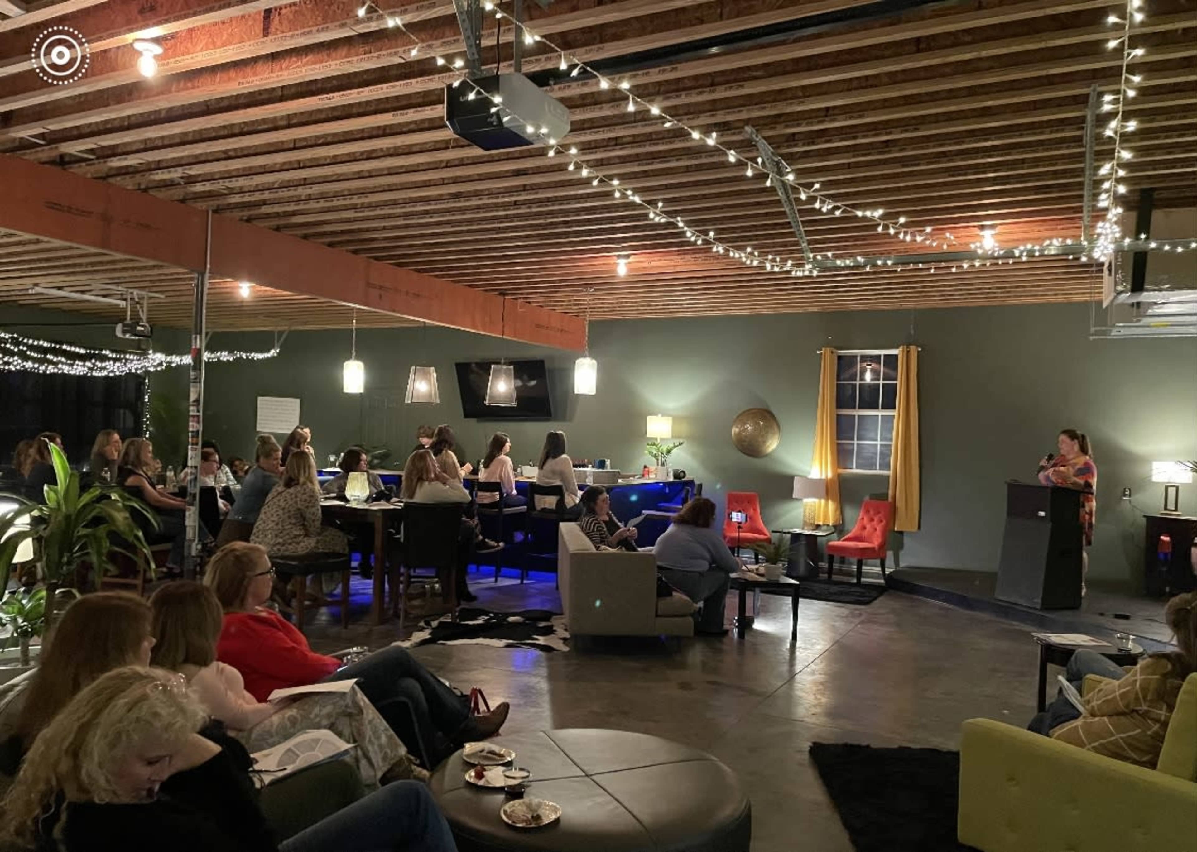 A group of people is seated in a stylish venue with string lights and modern decor, listening to a speaker at a podium.