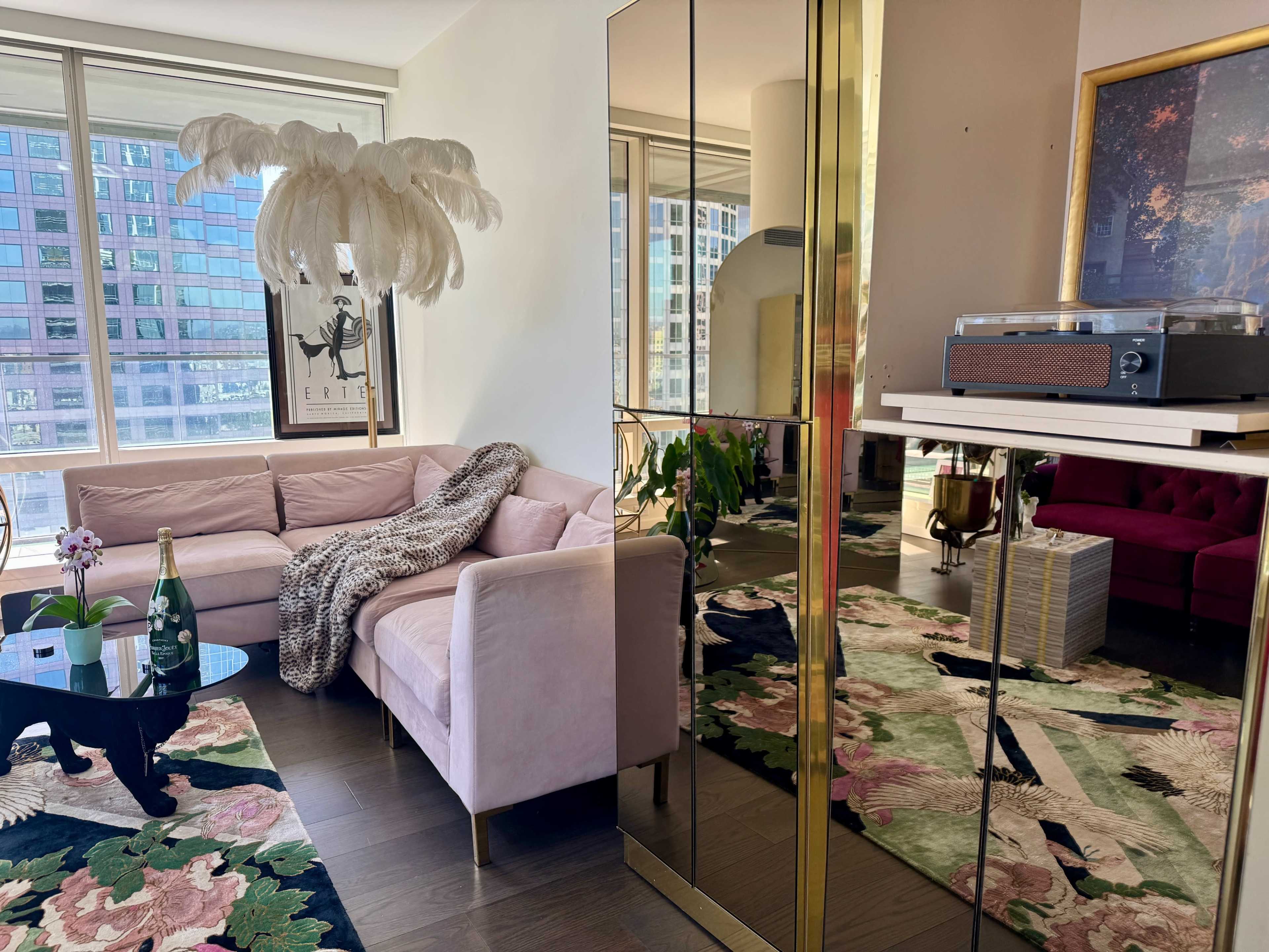 The image depicts a stylish living room featuring a pink couch, a glass coffee table with a plant, decorative mirrors, and a record player, with large windows offering a view of a cityscape.