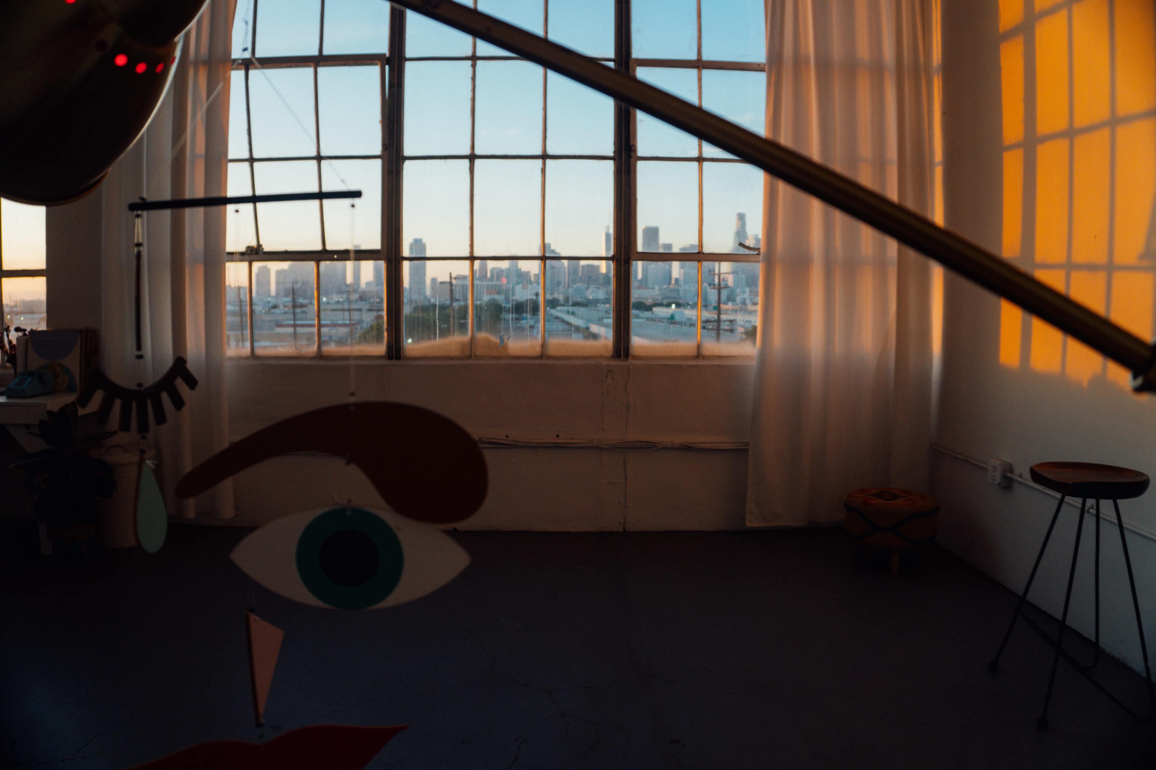 The image shows an urban studio with large windows revealing a city skyline at sunset, while a decorative mobile featuring abstract facial features hangs in the foreground.