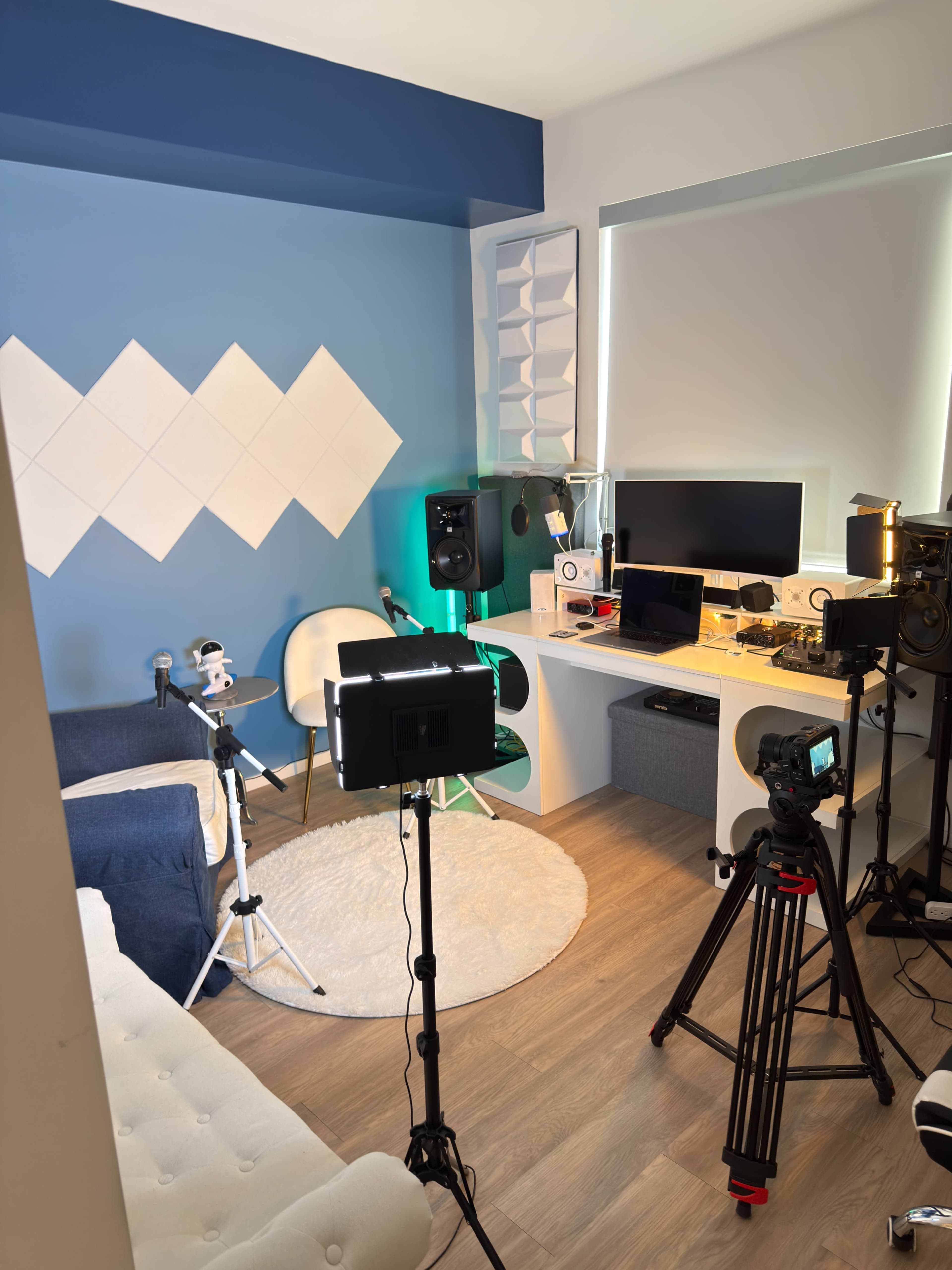 The image shows a brightly lit room arranged as a multimedia setup, featuring a white rug, a desk with various electronic devices, and multiple stands with lights and cameras aimed towards a seating area.