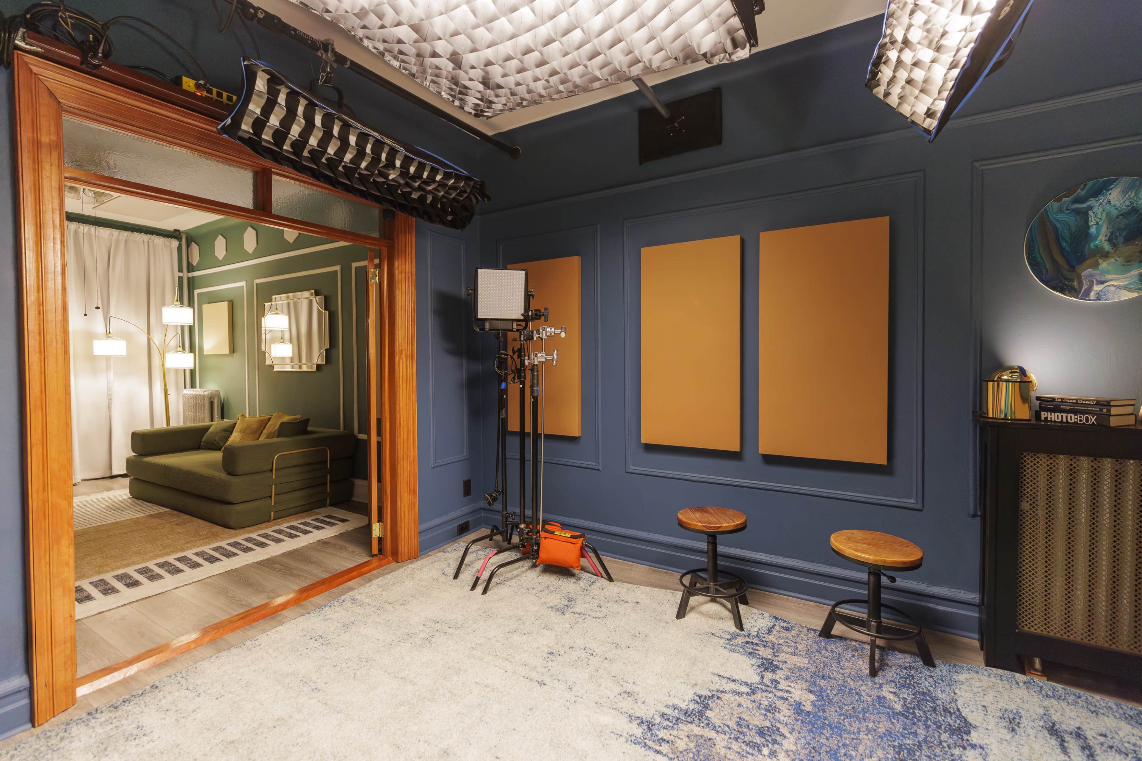 The scene shows a well-lit studio with a textured blue wall, two empty wooden stools, and a doorway leading to a room with a green sofa and hanging light fixtures.