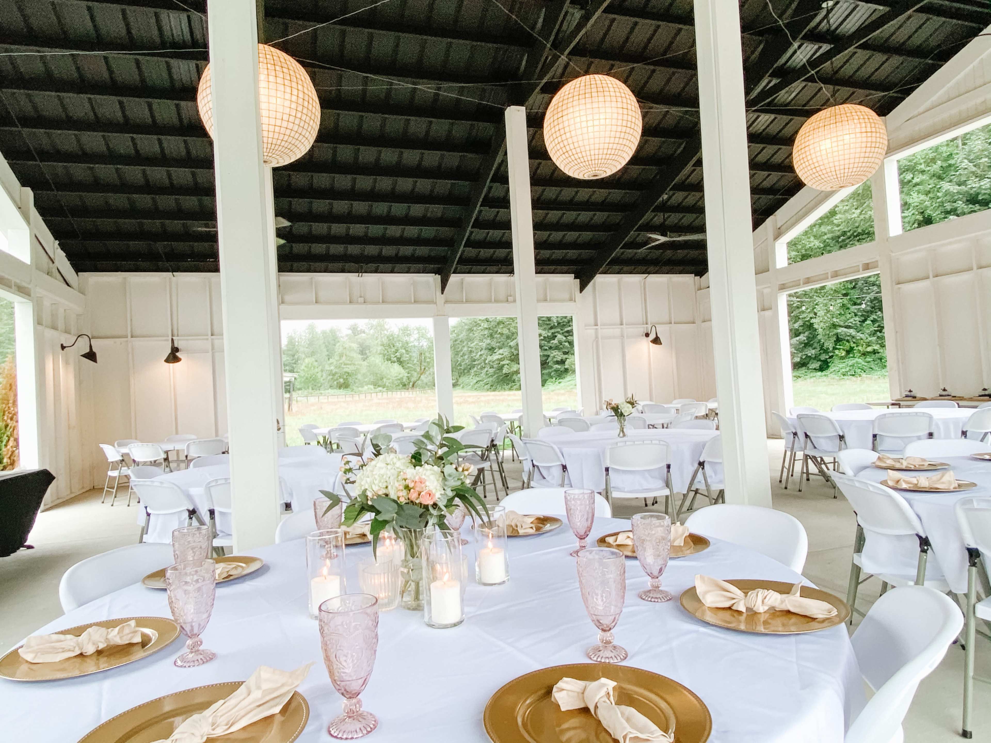 An indoor event space features white tables set with golden chargers, glassware, and floral centerpieces, illuminated by woven pendant lights.