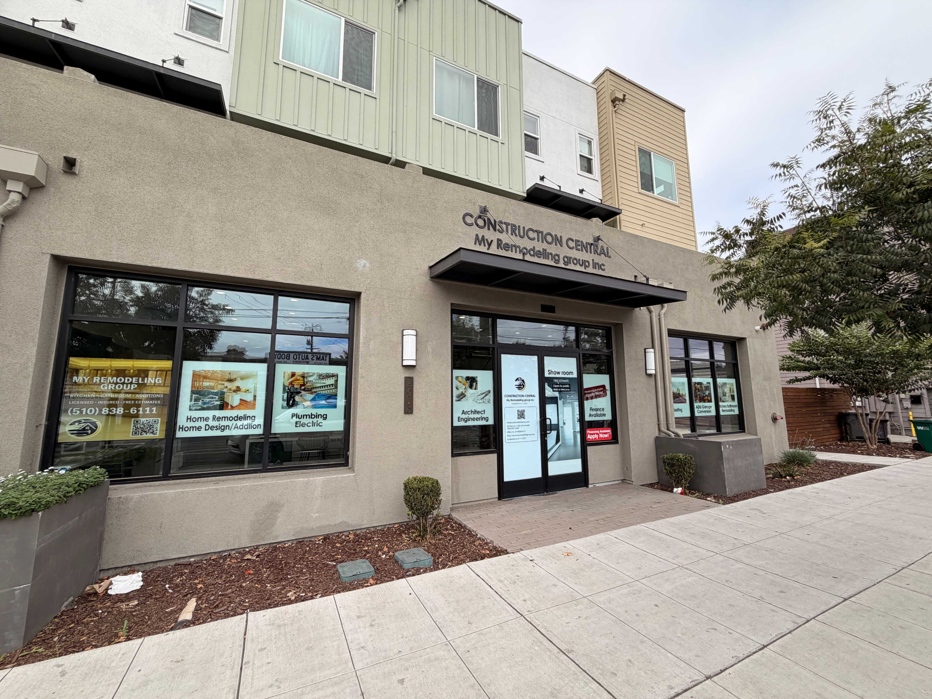 The image shows the exterior of a commercial building with a sign for "My Remodeling Group Inc," featuring large windows displaying promotional materials and a landscaped walkway.