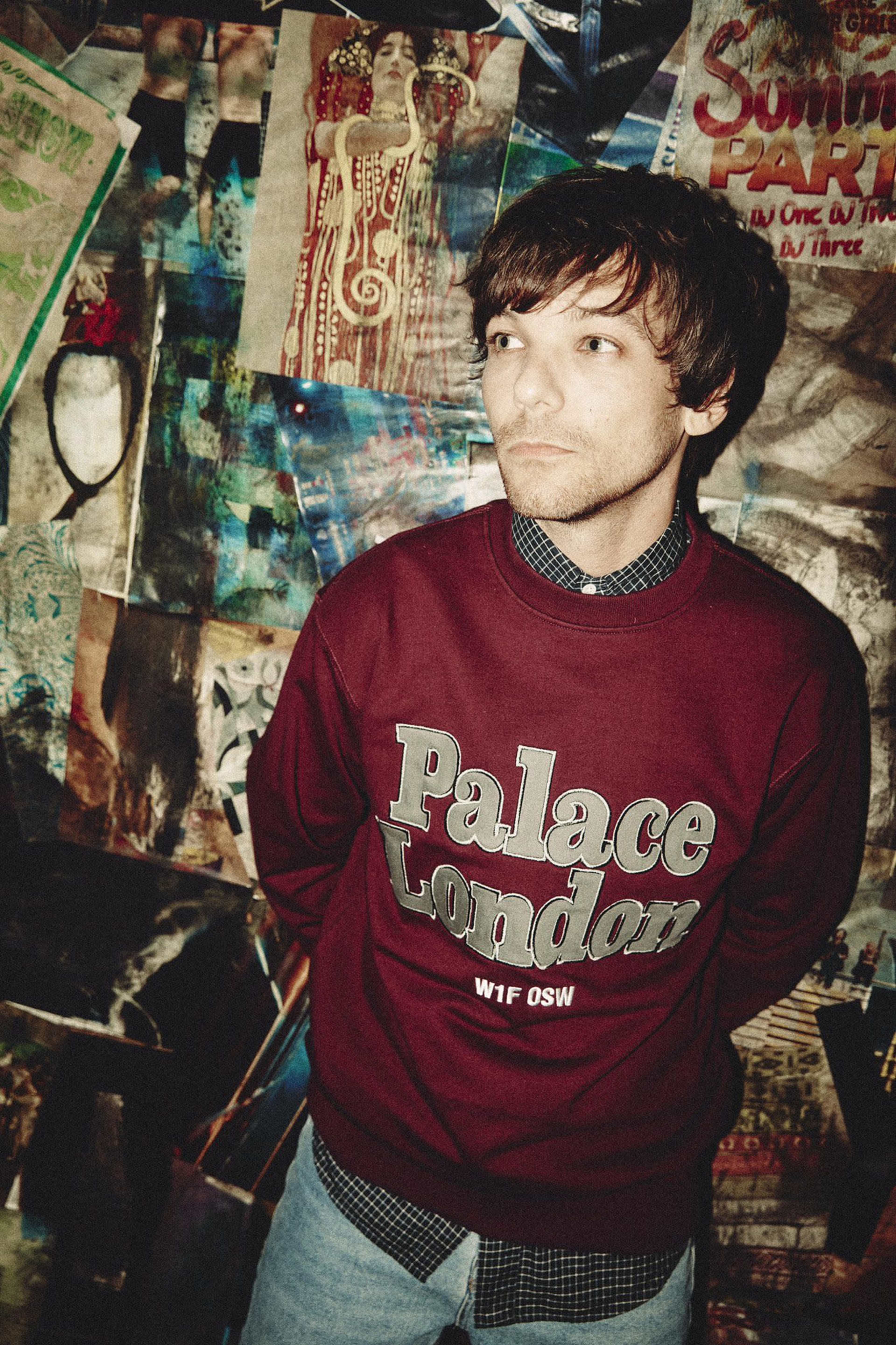 A man stands against a wall covered with various posters and artwork, wearing a maroon sweatshirt with "Palace London" printed on it.