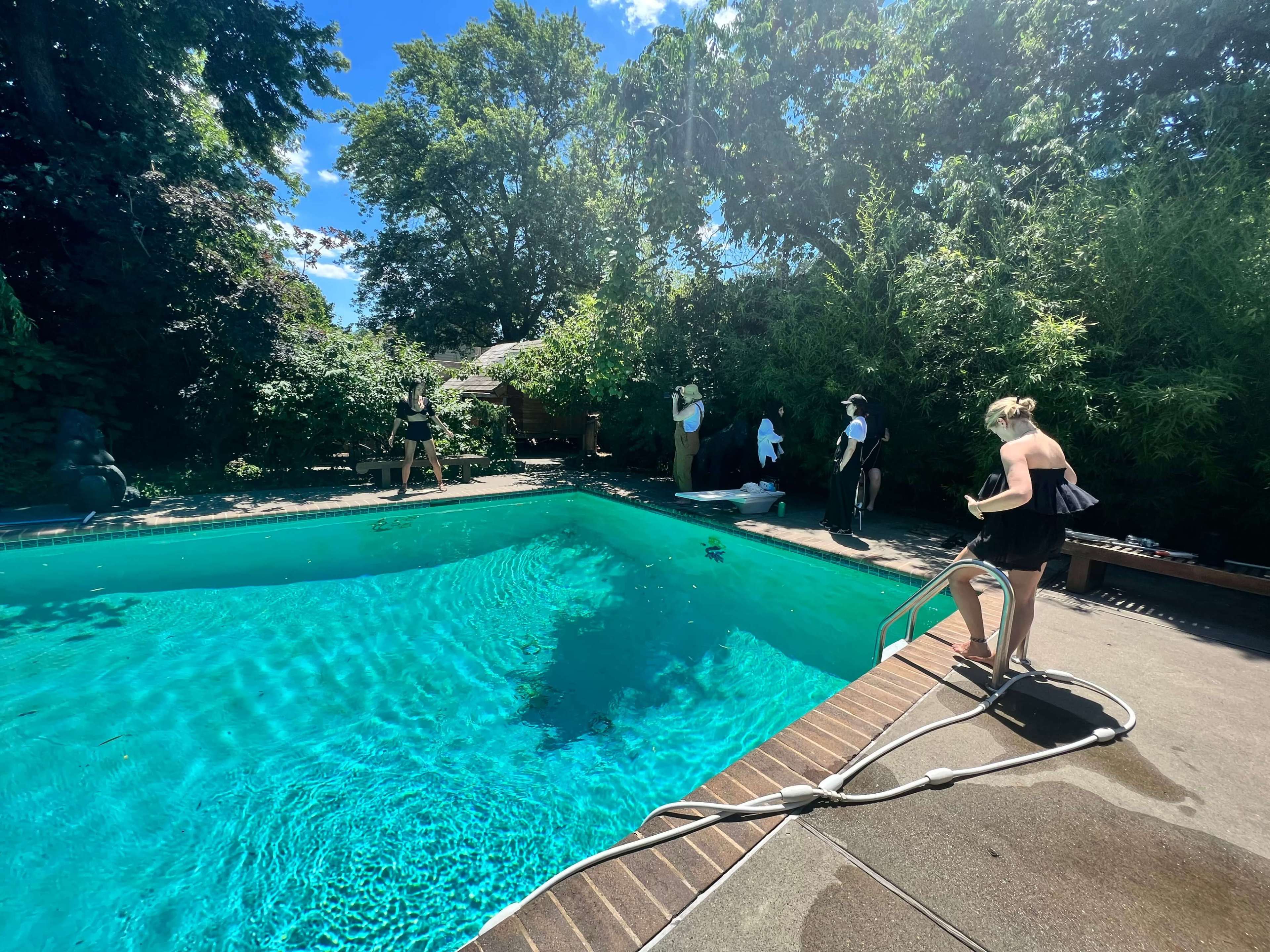 A swimming pool surrounded by greenery, with a few people near the edge and one person preparing to enter the water.