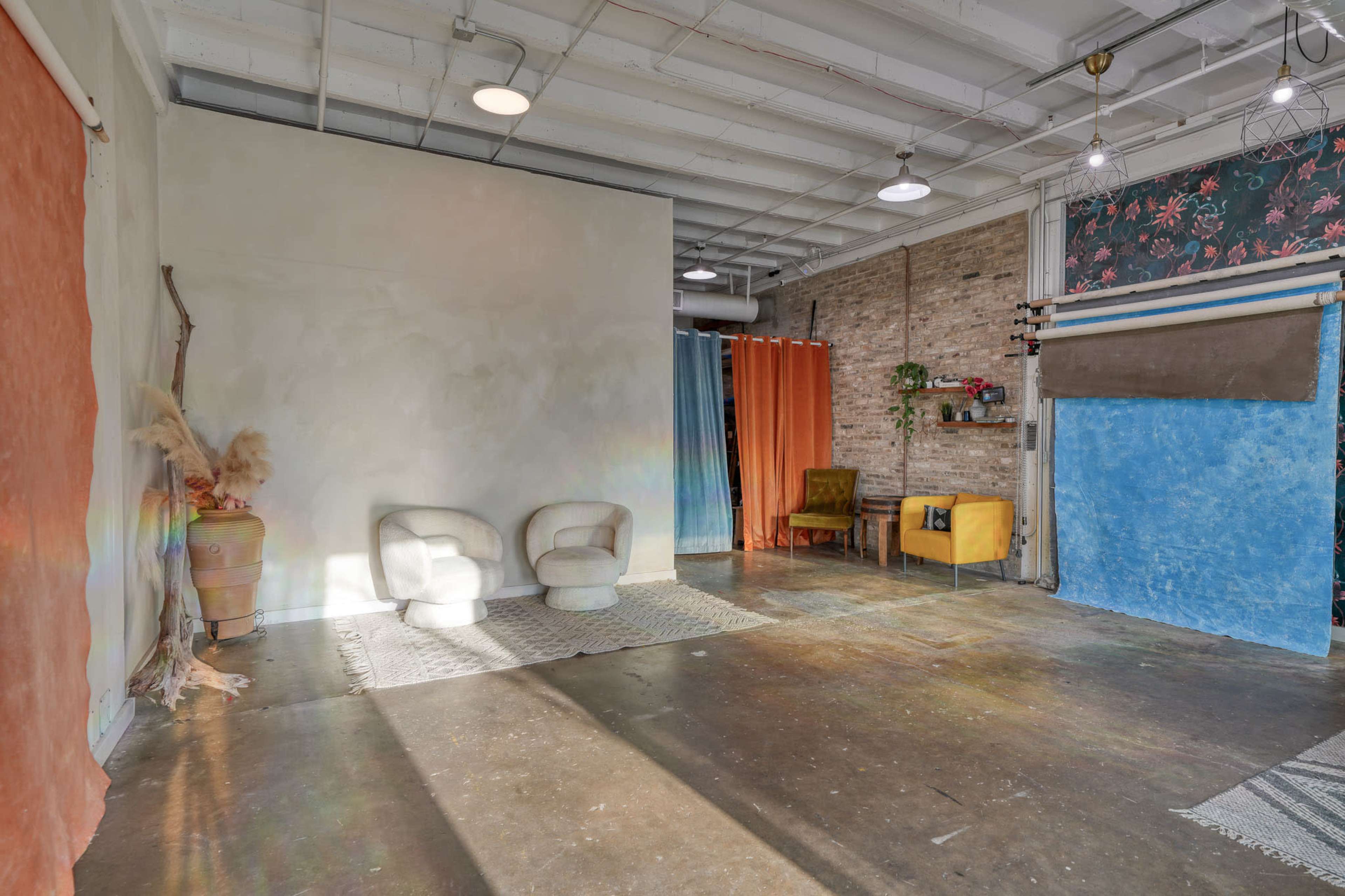 The image shows a spacious studio with an eclectic arrangement of colorful backdrops, two chairs in the foreground, and natural light illuminating the floor.