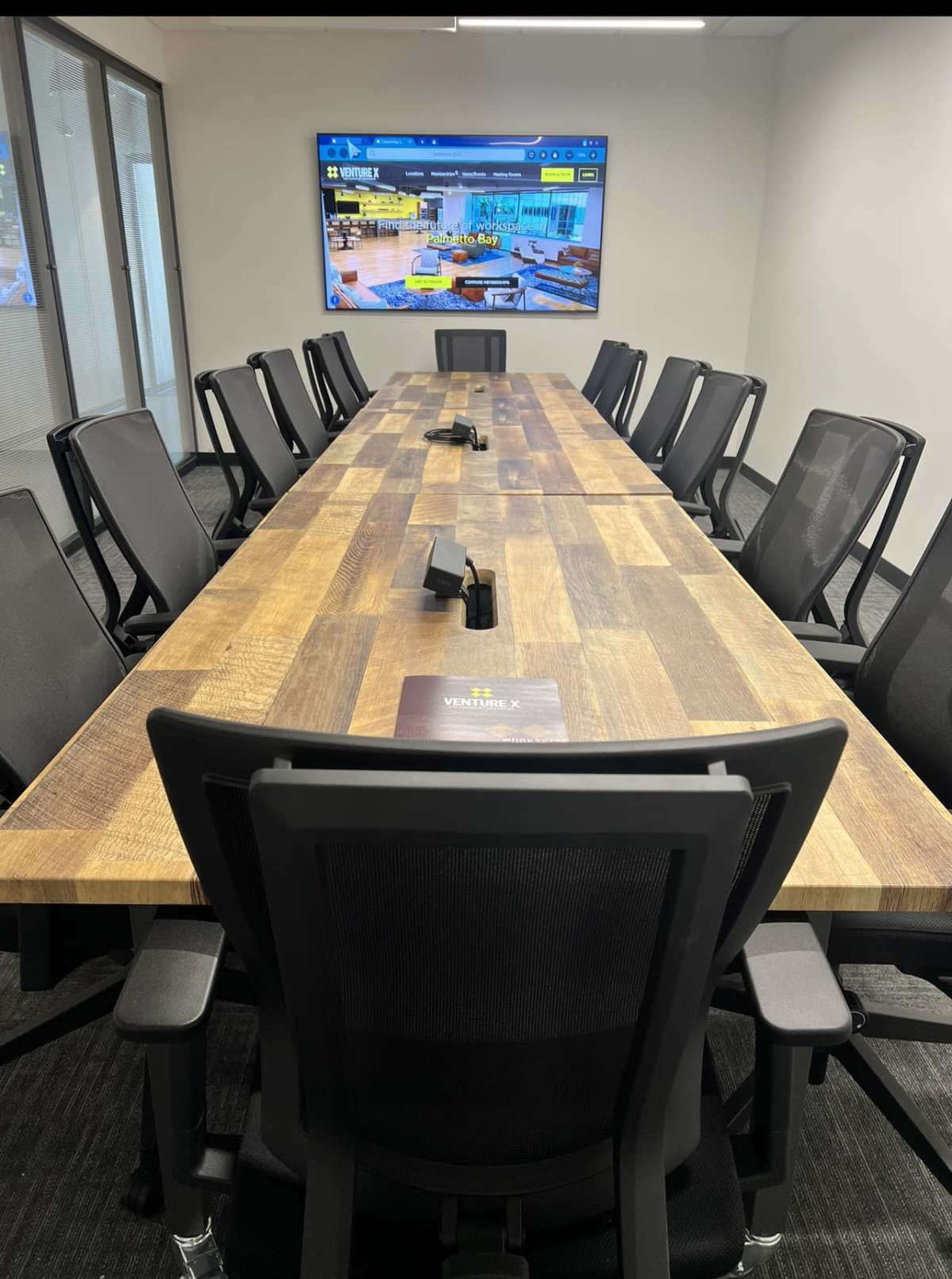 A long, wooden conference table with multiple black chairs is set up in a meeting room that features a large screen on one wall.