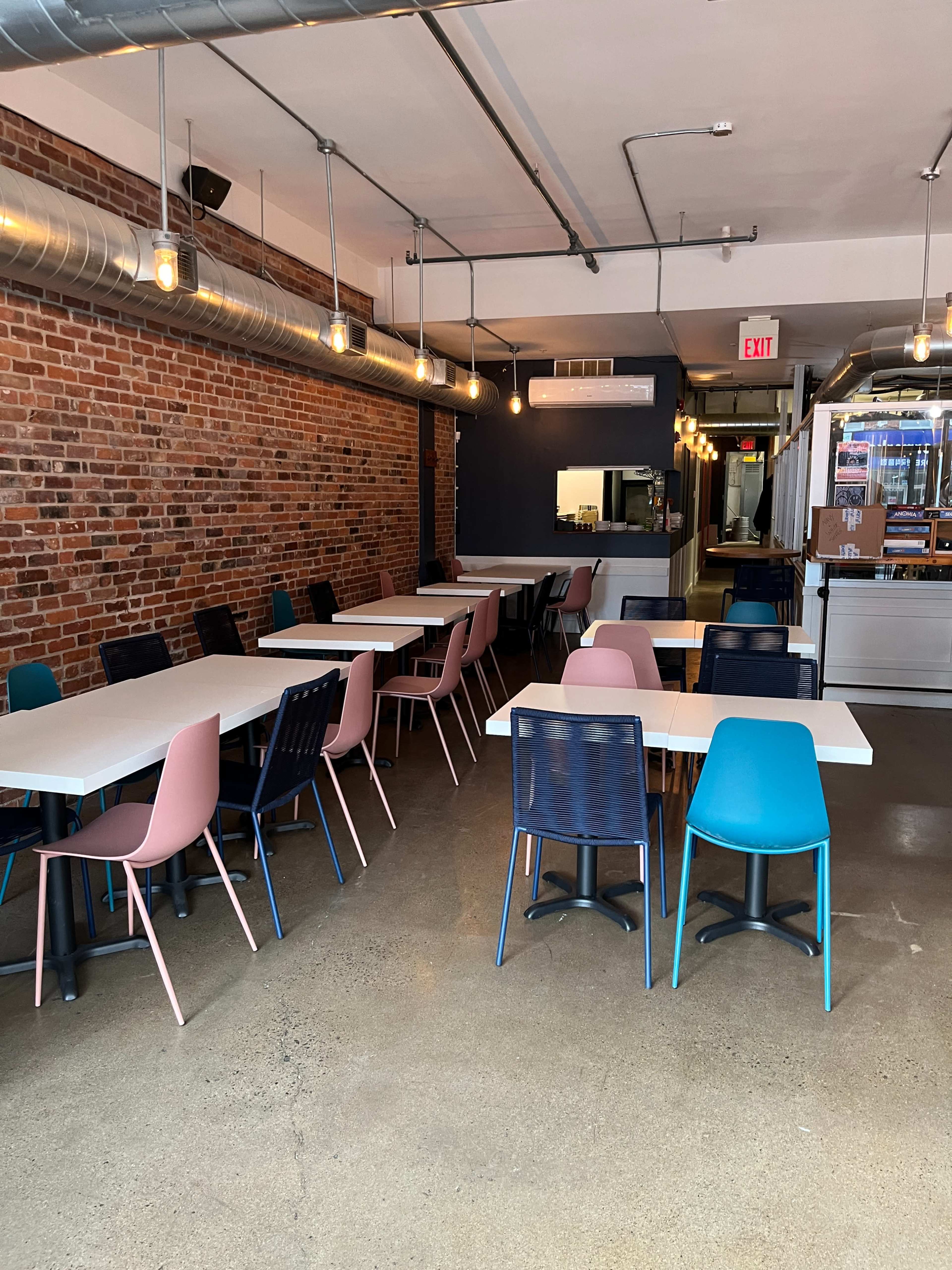 A spacious cafe with a mix of light pink, blue, and dark chairs arranged around white tables against a backdrop of exposed brick and industrial-style lighting.