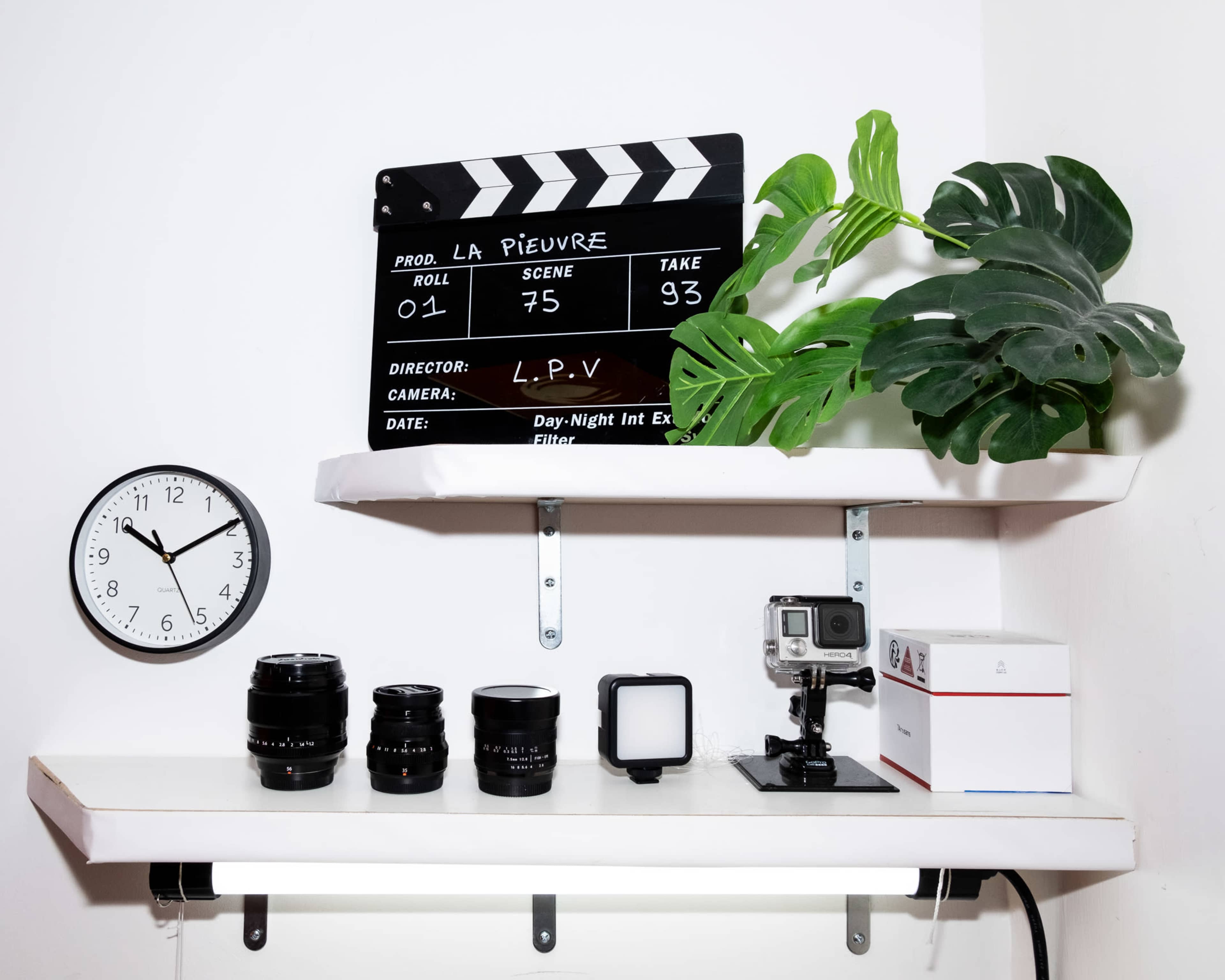 A film clapperboard, camera lenses, a clock, and a GoPro camera are arranged on two white shelves with a green plant in the background.