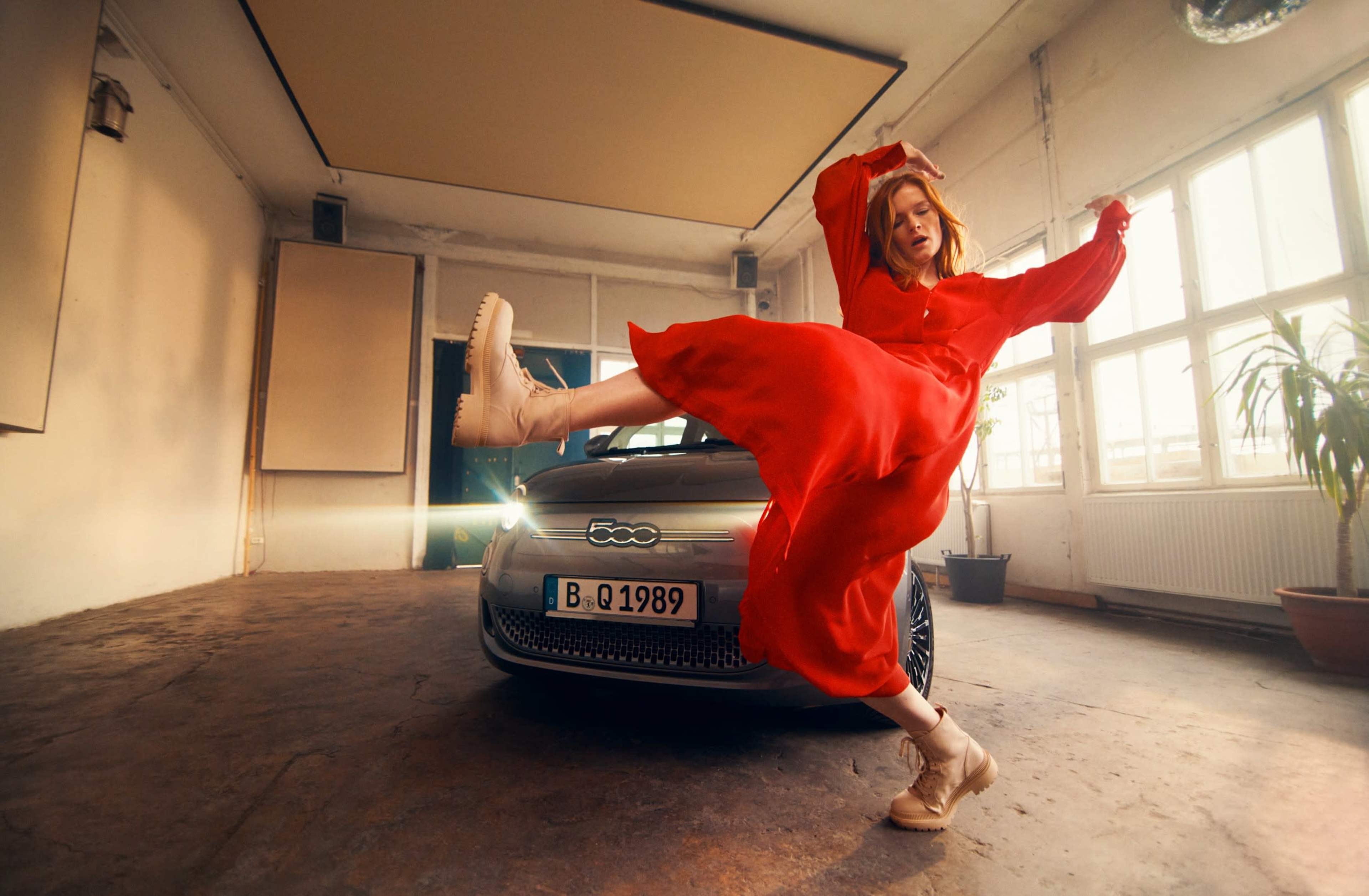 A person in a red dress poses dynamically next to a silver car in a brightly lit interior space.