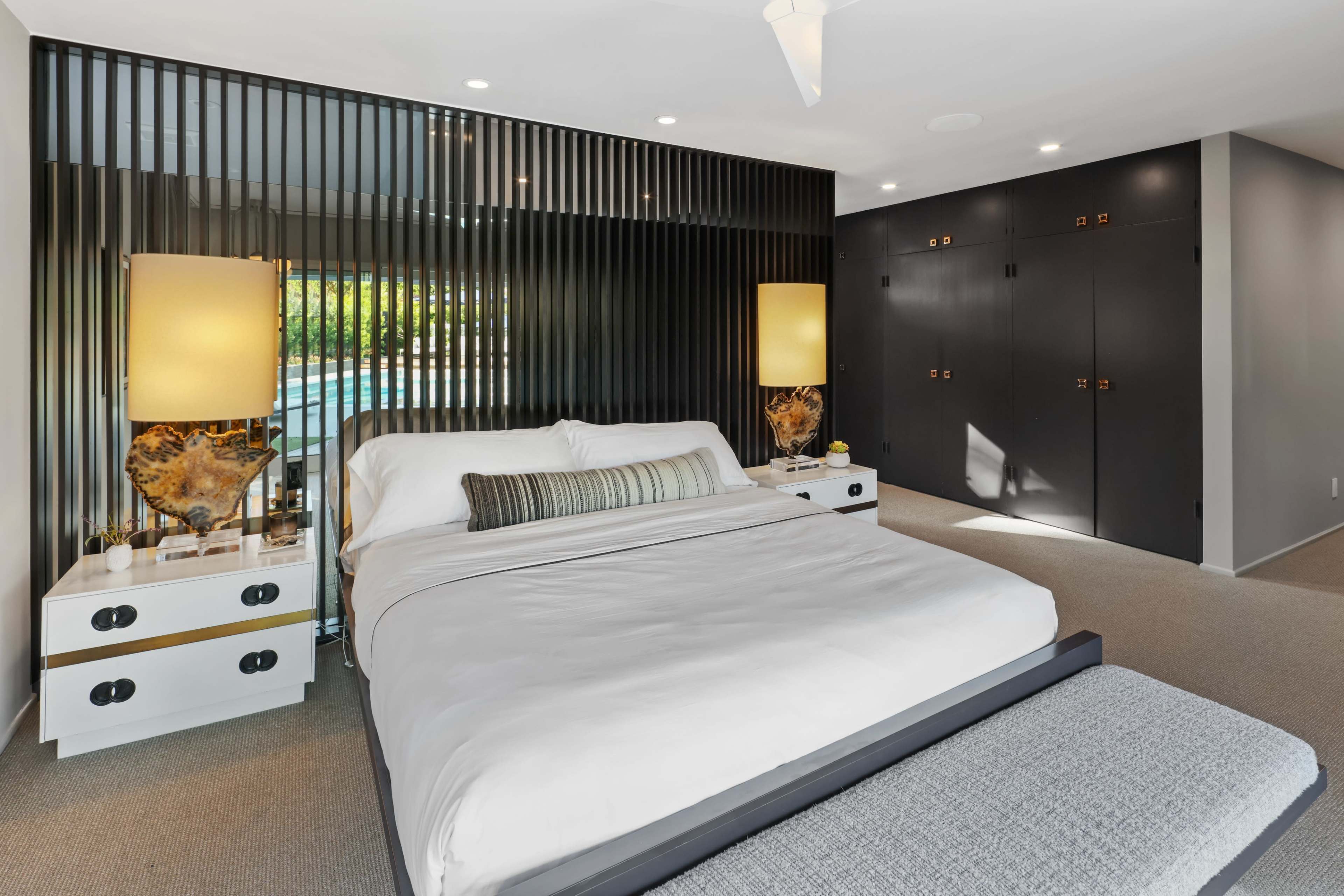 The image depicts a modern bedroom featuring a large bed, two bedside tables with lamps, and a wall with vertical slats behind the bed, alongside dark cabinetry.