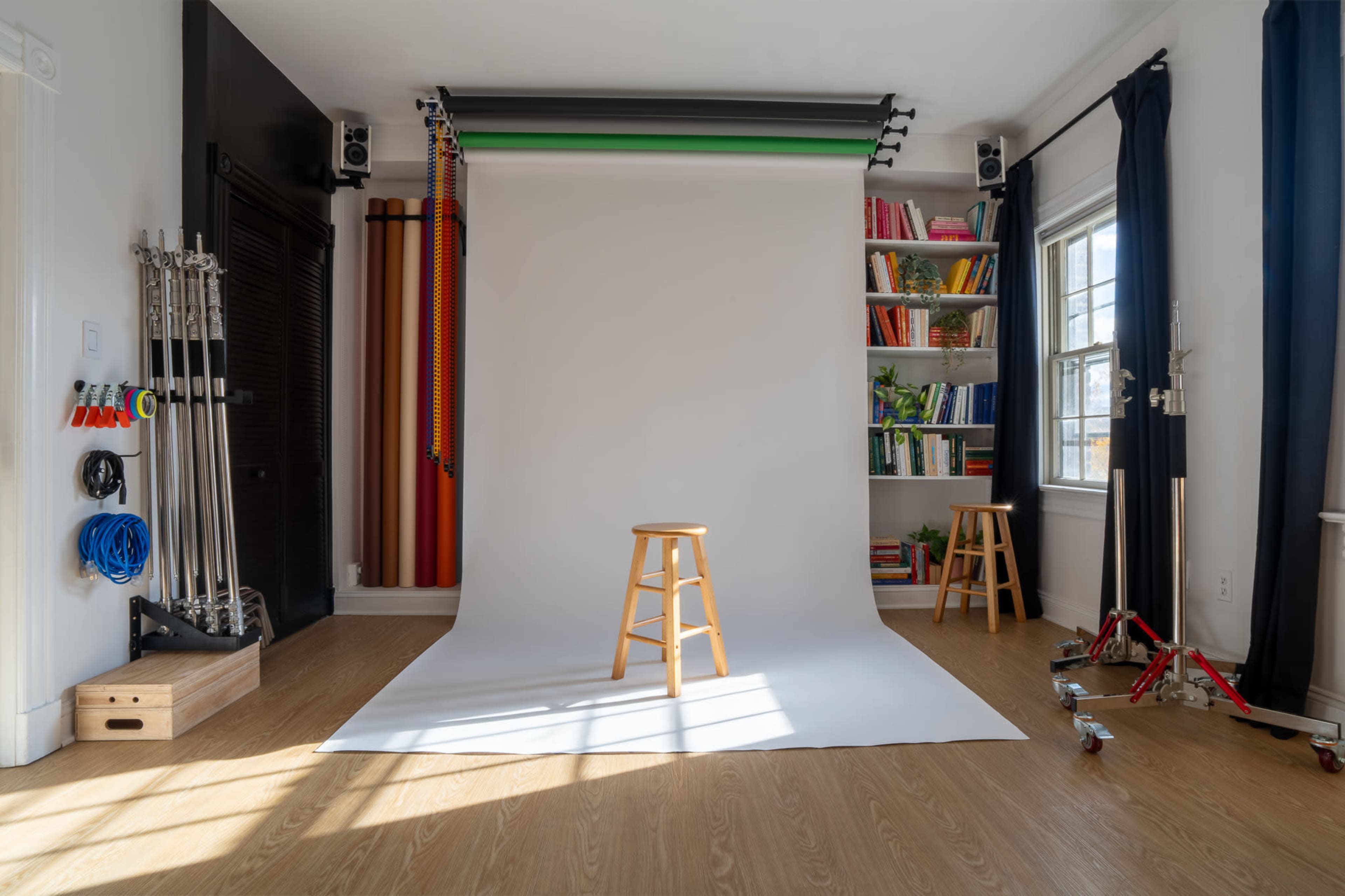 The image shows a photography studio with a backdrop, a stool in the center, and colorful backdrops hanging from the ceiling, along with bookshelves filled with books on one side.